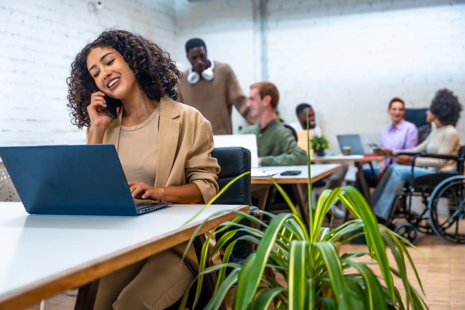 Woman talking to the mobile phone and using laptop in a coworking space with coworkers on the background