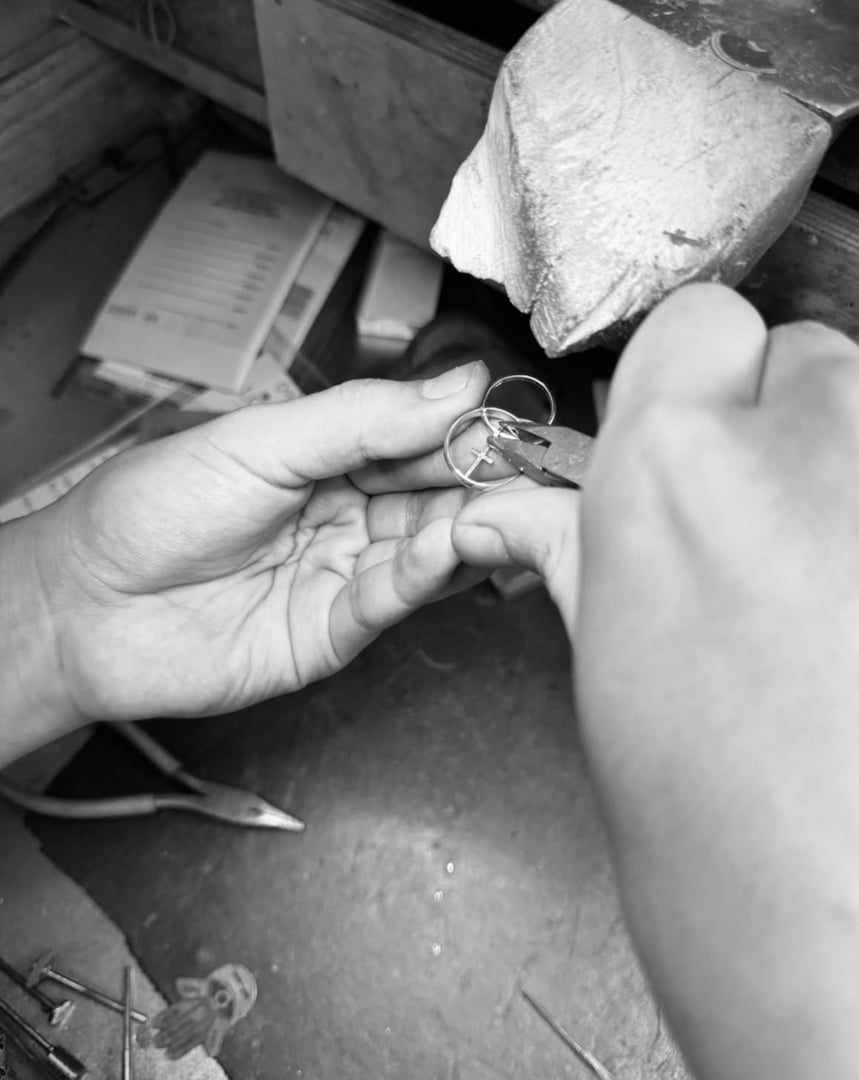 Hands crafting jewelry with pliers at a workbench, surrounded by tools and notes. Monochrome image.