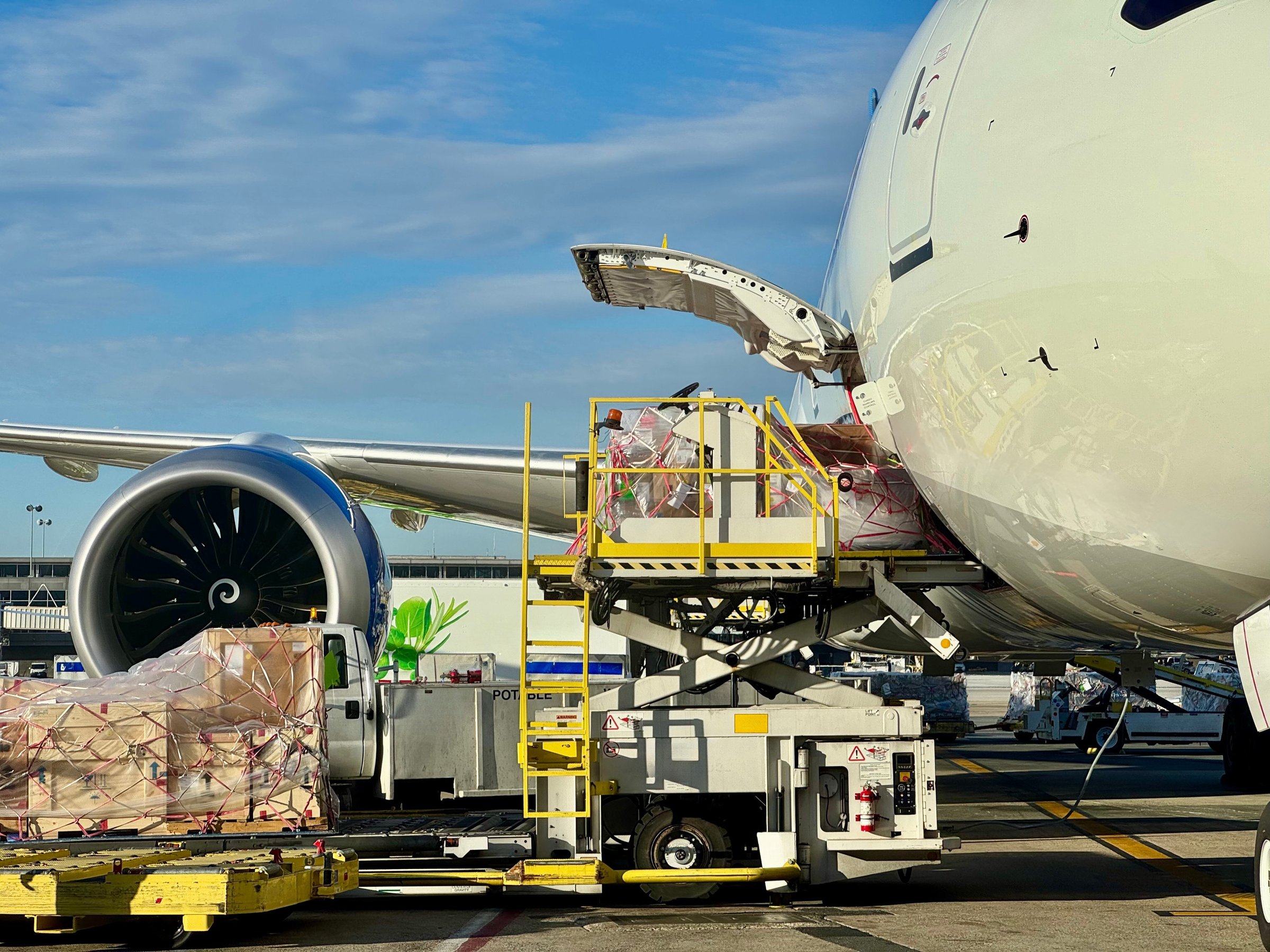 Cargo operations at a commercial airport. The cargo door is open and heavy cargo is unloaded onto a lift vehicle. Crates are stacked under a cargo net on a pallet.  The shiny metal skin of the airplane reflects the operation.