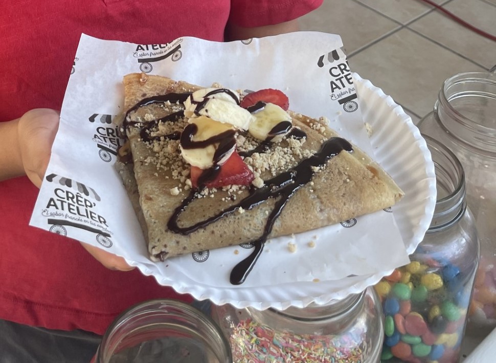 Crepe with ice cream, strawberries, nuts, and chocolate drizzle on a paper plate, held by a person.