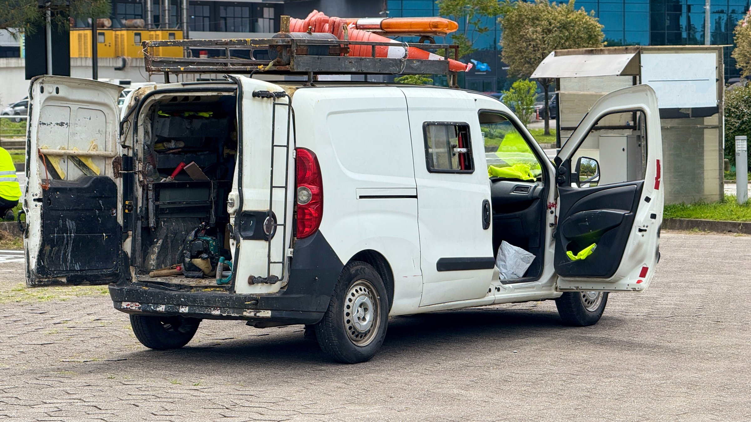 This image features a white utility van parked outdoors, with both the rear and side doors open, revealing an array of tools and equipment. Inside the van, a toolbox, power tools, and various other items are organized for easy access. On the roof, traffic cones and additional work gear are secured within a metal rack. Nearby, individuals wearing high-visibility jackets are present, suggesting an active maintenance or construction project. The background includes greenery and buildings, further establishing the urban or industrial setting. This scene is ideal for themes related to professional work vehicles, maintenance crews, or outdoor service operations.