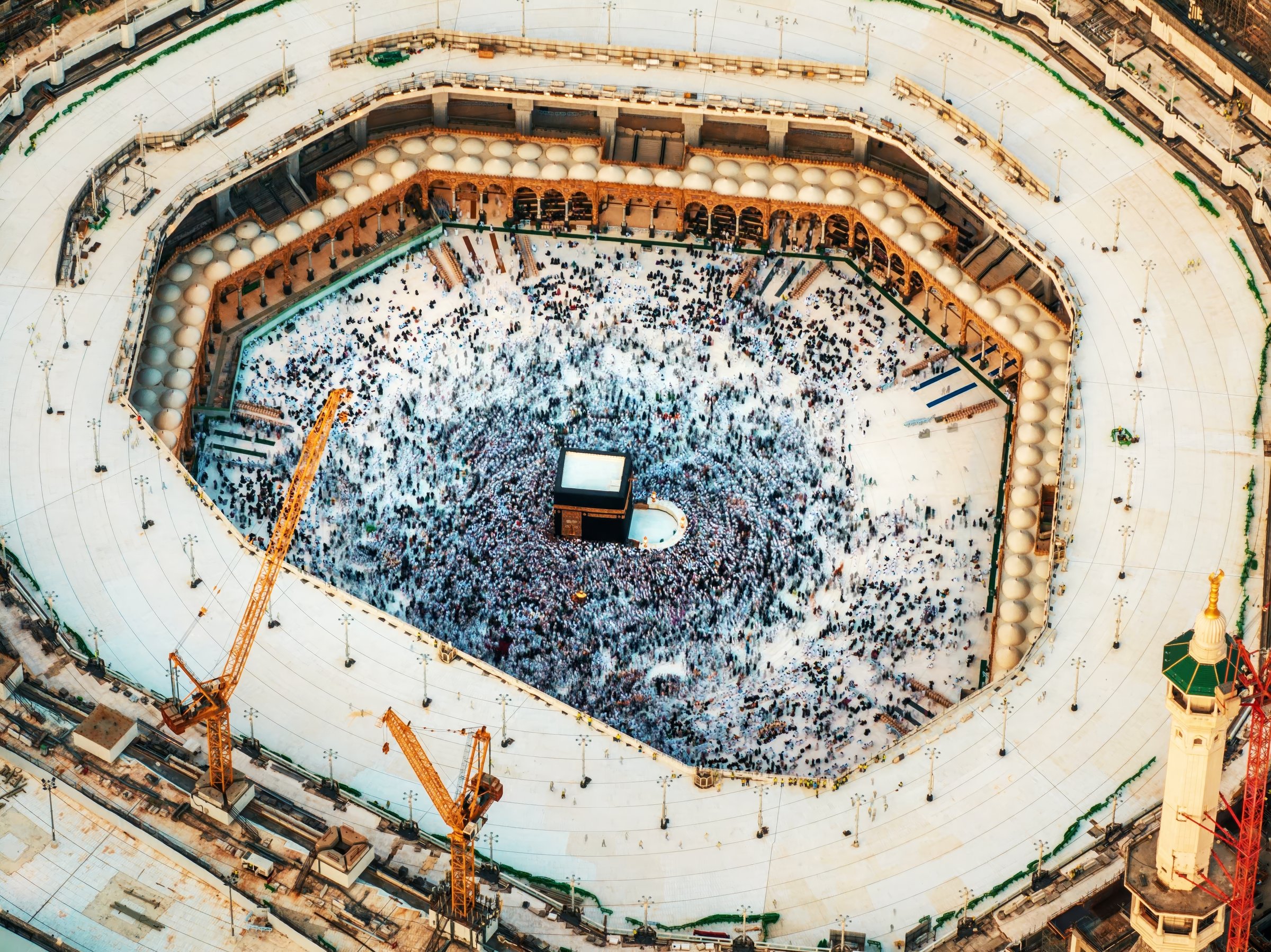 Aerial view showing crowds of pilgrims gathered around the Kaaba in the Grand Mosque, with construction cranes surrounding the site in Mecca