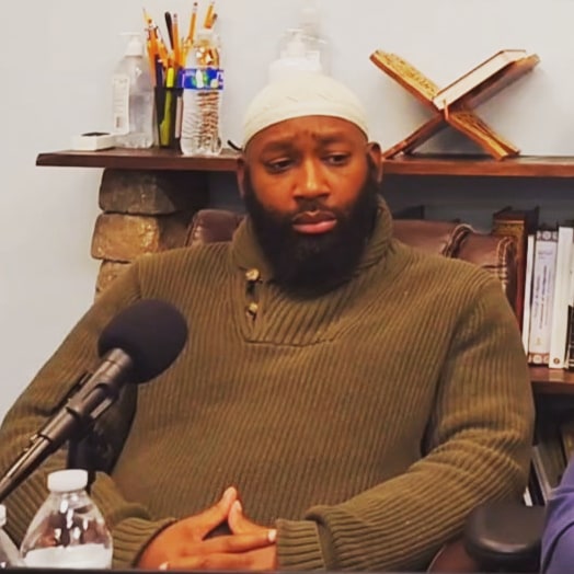 A man with a beard wearing a green sweater sits at a table with a microphone, books, and a water bottle nearby.