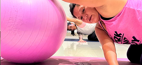Two women stretching on exercise mats with a pink stability ball in the foreground, both wearing workout attire.