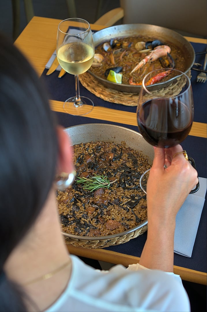 Over-the-shoulder shot of a person enjoying a gourmet paella meal with a glass of red wine at a restaurant. The table is elegantly set with another glass of white wine and a second pan of seafood paella. This image is ideal for culinary blogs, restaurant advertisements, and food industry promotions, showcasing a luxurious dining experience.