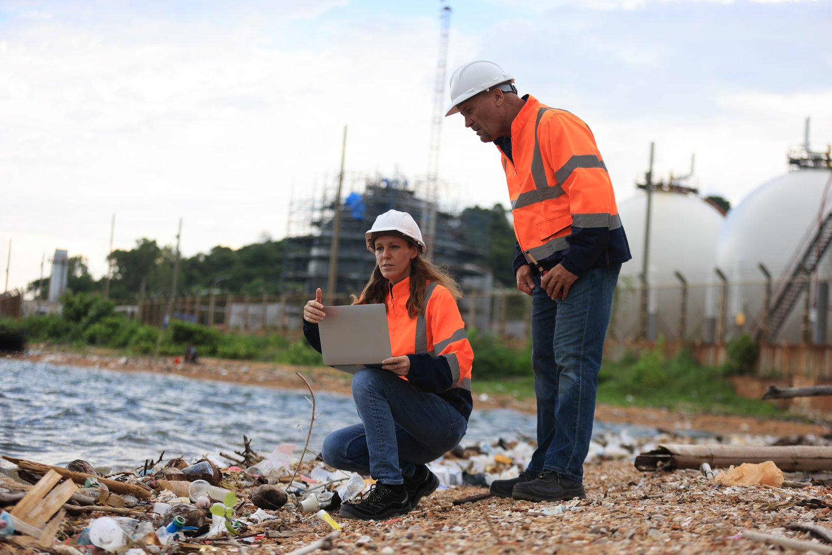 A seasoned female Chief Petrochemical Engineer and a senior male Engineer are confidently discussing and analyzing the production performance of oil and gas while standing side by side and wearing safety coveralls. They are using a laptop to conduct their research on the impact of wastewater within the industry area.
