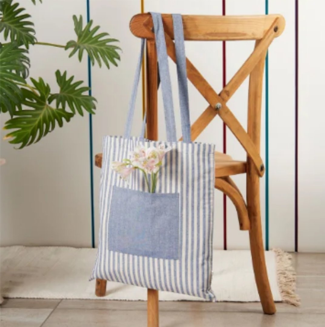 Blue striped tote bag with flowers in the pocket hanging on a wooden chair next to a plant.