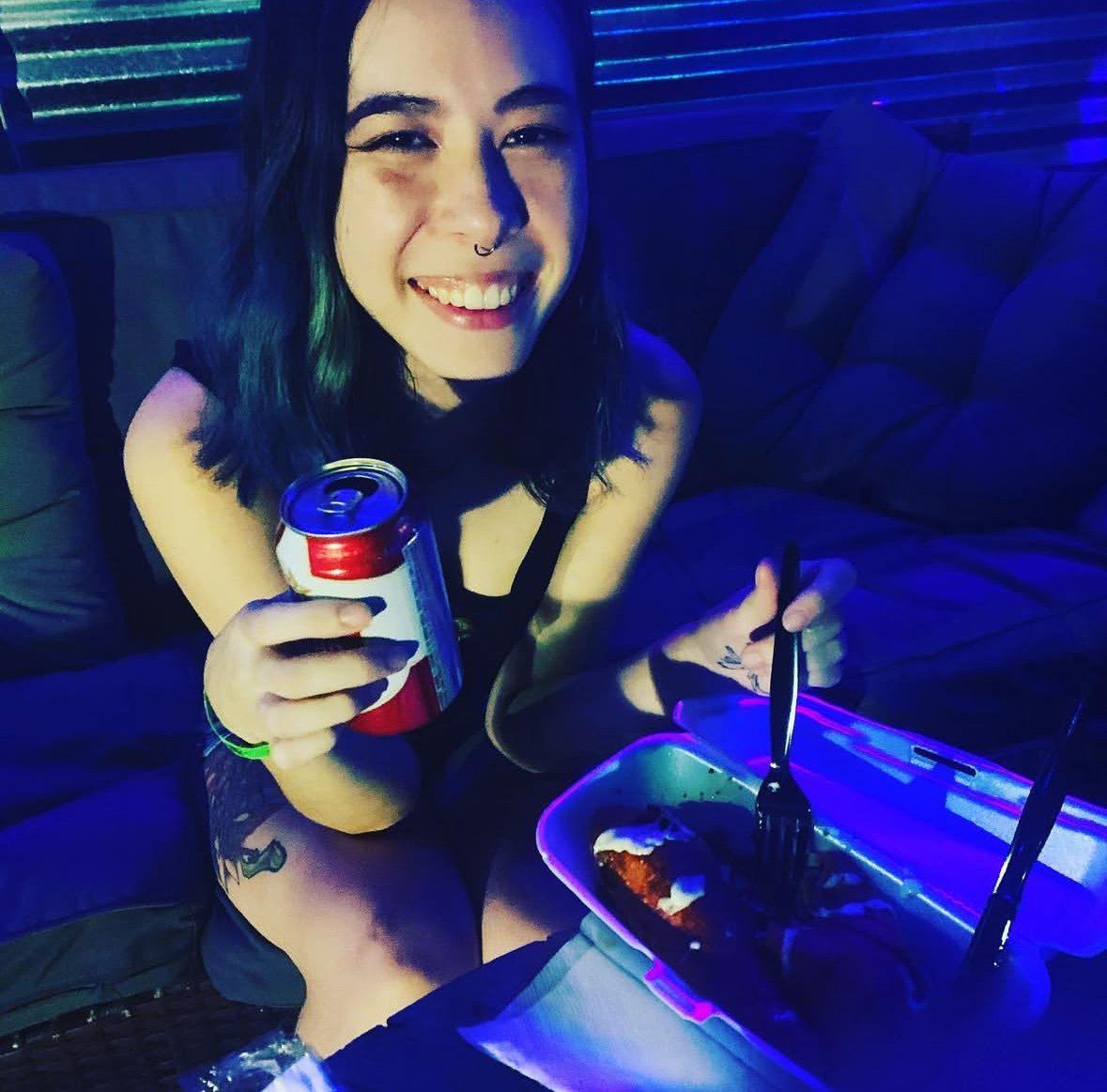 Smiling person holding a can and eating from a takeout container while sitting on a sofa in a dimly lit room.