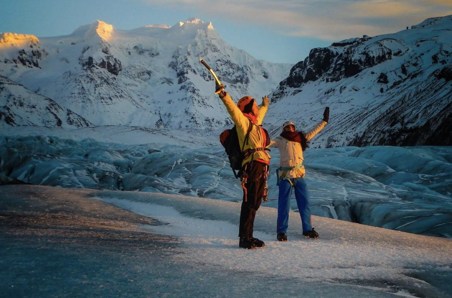 Two hikers with ice axes celebrate on a snowy glacier with mountains in the background at sunset.