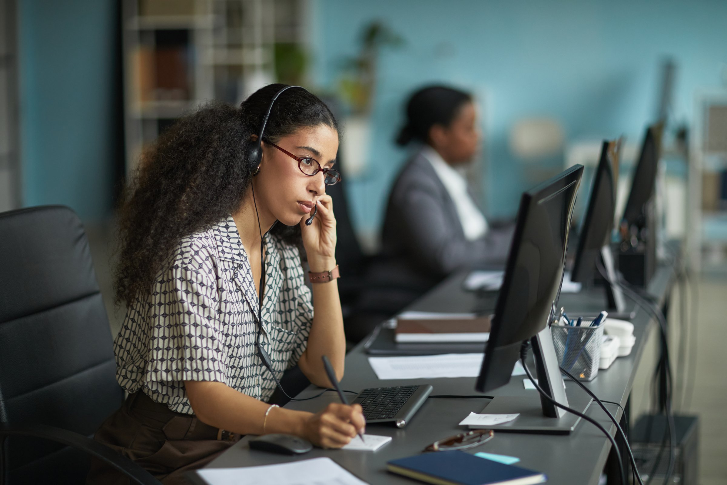 Young adult biracial woman wearing headset working at computer in modern office, holding pen and listening attentively, another Black woman working in background