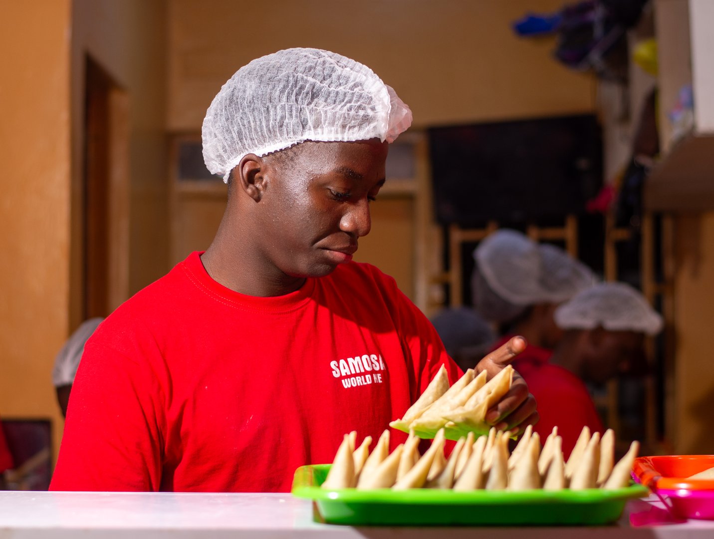 Chef in a red shirt and hairnet prepares samosas in a kitchen with trays of folded pastries on the counter.