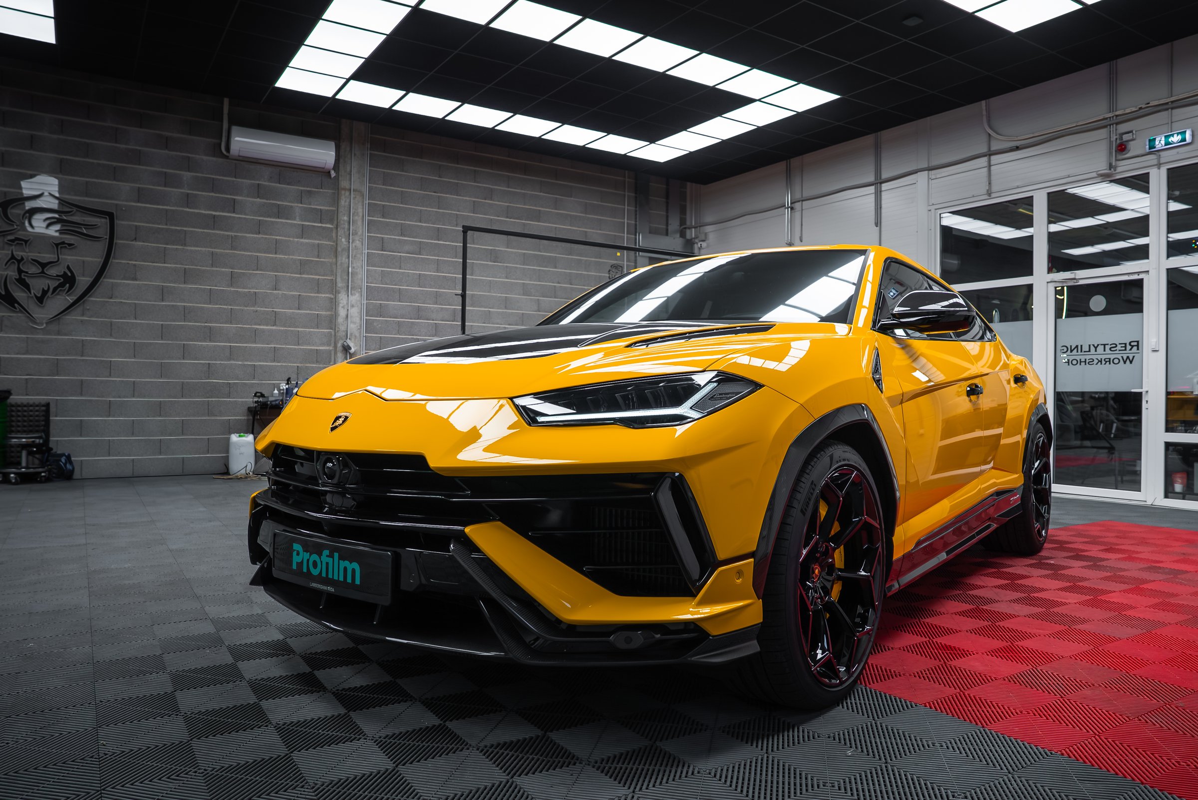 Yellow Lamborghini Urus Performante sits on a black and red checkered garage floor, low angle view shows aggressive front, black wheels, orange calipers, and carbon fiber accents.