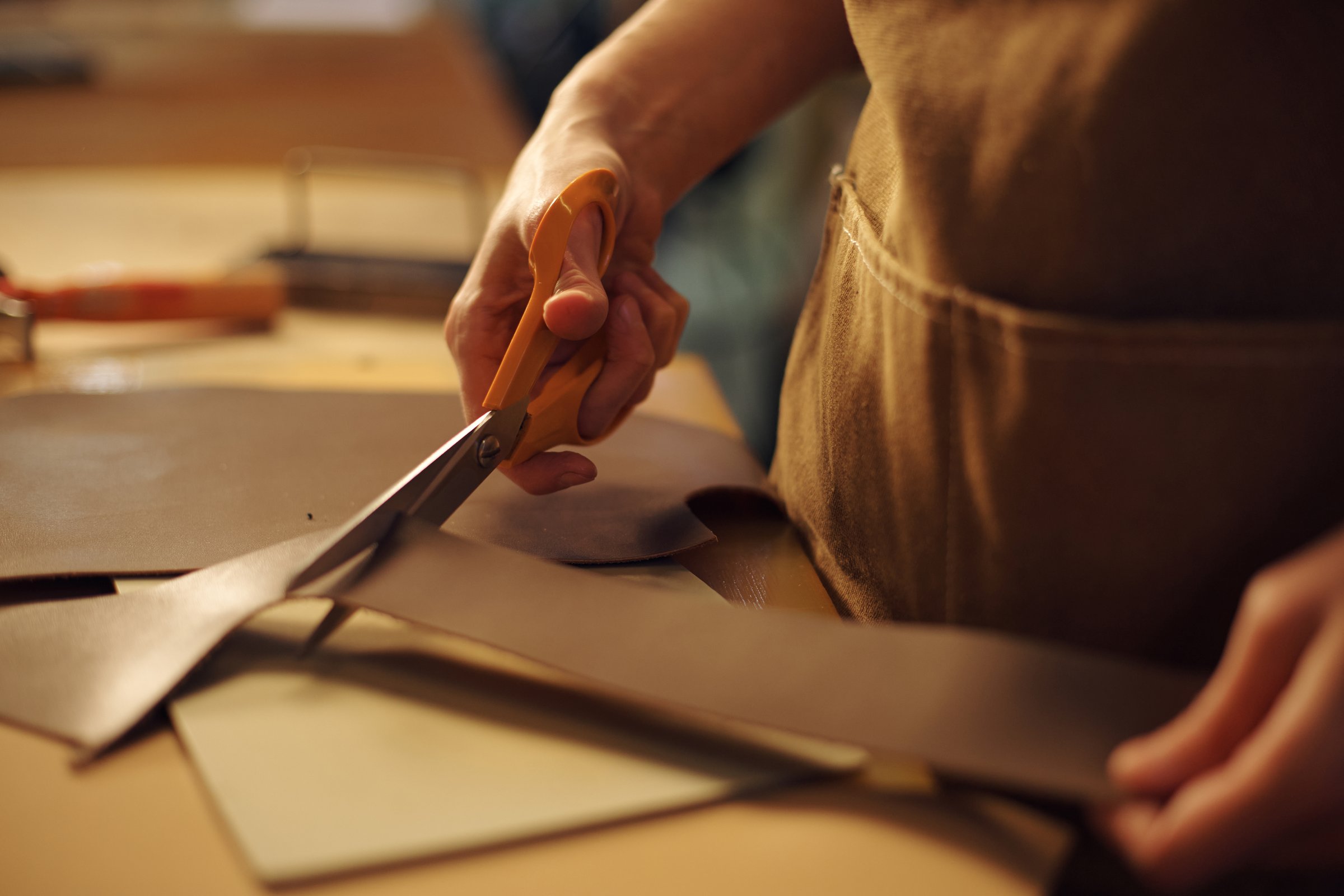 Close-up of craftsman's hands holding scissors and precisely cutting leather material in workshop environment focused on intricate craftsmanship and attention to detail