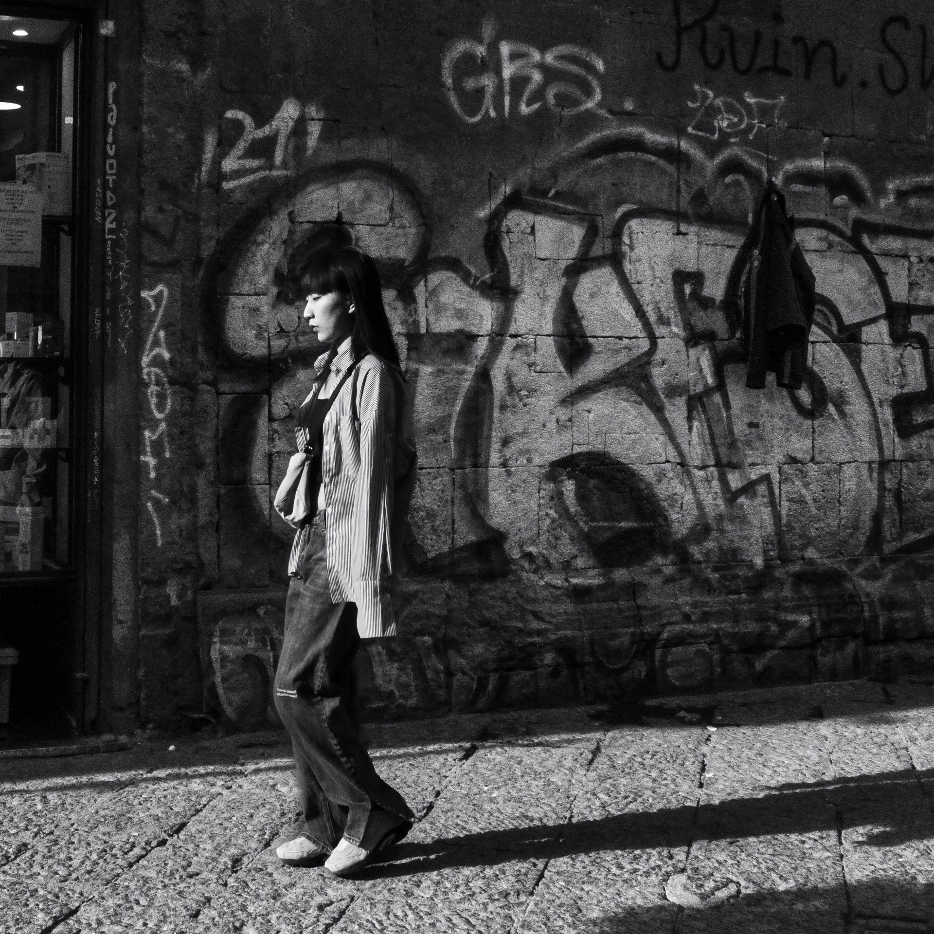 Black and white editorial photograph of a young girl walking along Via Toledo in Naples. The image captures the city's daily life. A realistic, spontaneous, and dynamic urban shot.