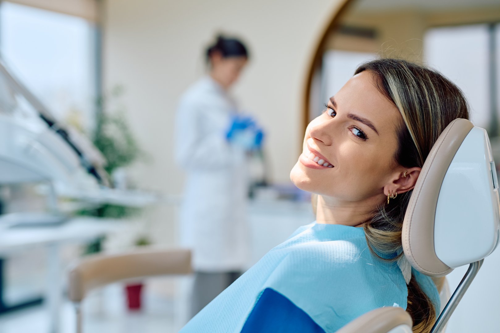 Young woman confidently smiling, sitting in a dental chair, a healthcare professional in uniform working in the background
