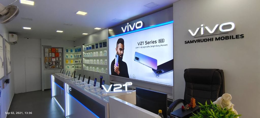 Interior of a Vivo mobile store displaying phones and promotional banners for V21 series on a well-lit showroom counter.