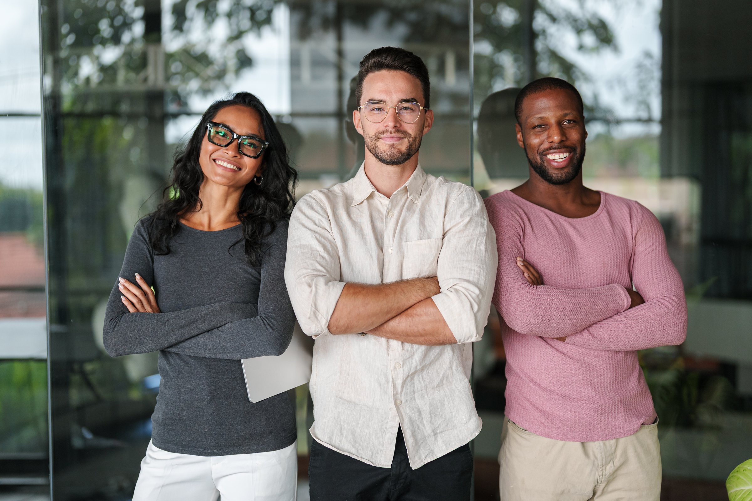 Portrait shot of three young professionals - Asian woman in glasses, Caucasian man with beard, and African American man - standing with crossed arms in modern office environment.