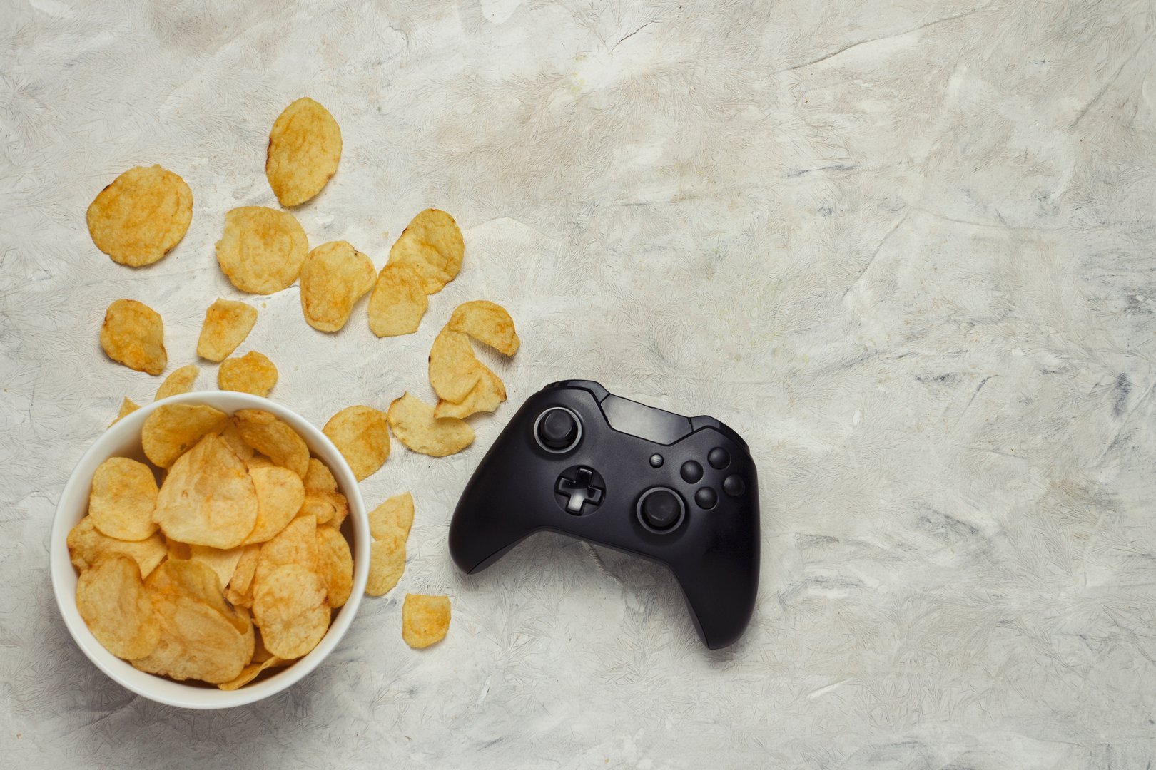 Gamepad and Bowl with Chips on a Light Stone Background. The concept of computer games and sports tournaments. Flat lay, top view.