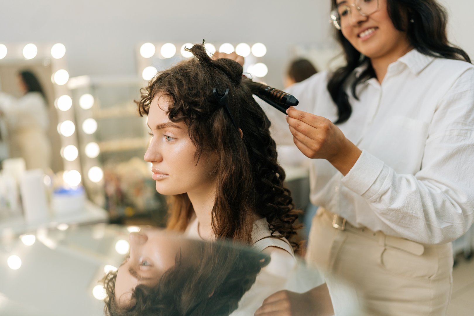 Smiling female hairstylist using hair iron and comb for young woman hairstyling in modern hairdressing studio. Hairdresser creating female hairdo with tongs. Concept of fashion and beauty.
