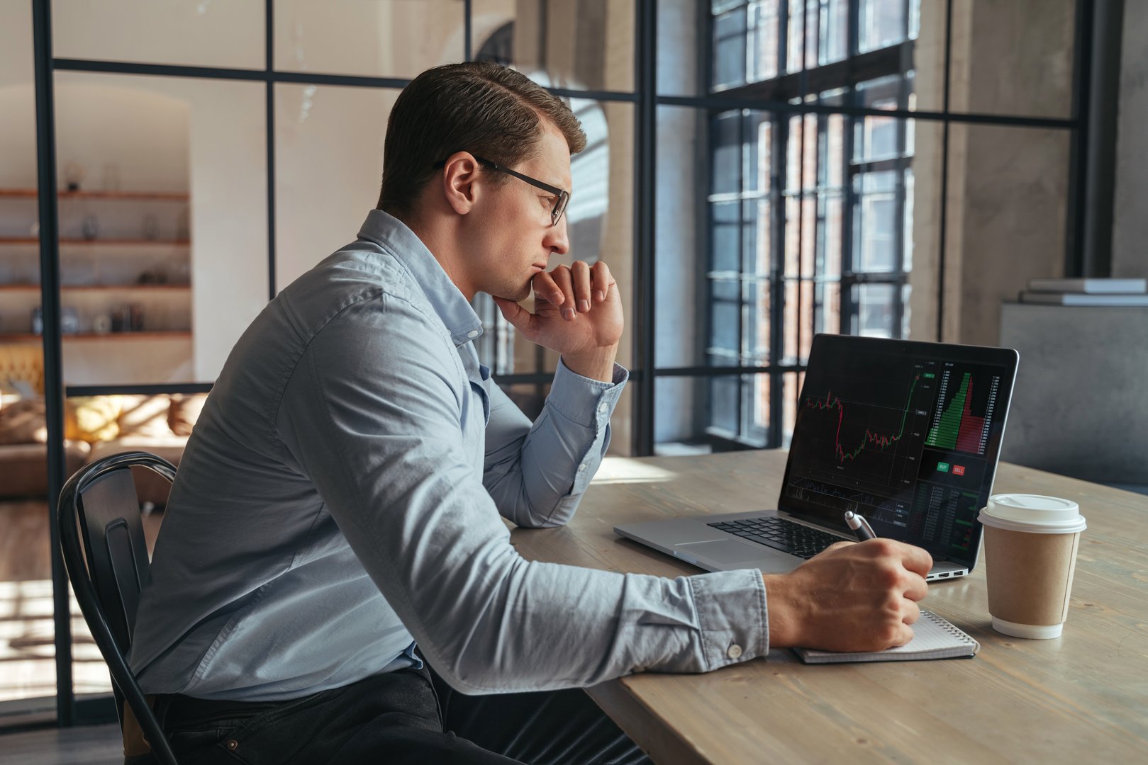 Serious crypto trader looking at laptop screen with chart, analyzing risks of buying cryptocurrency in the conditions of changing world and global recession, sitting at his office table, making notes