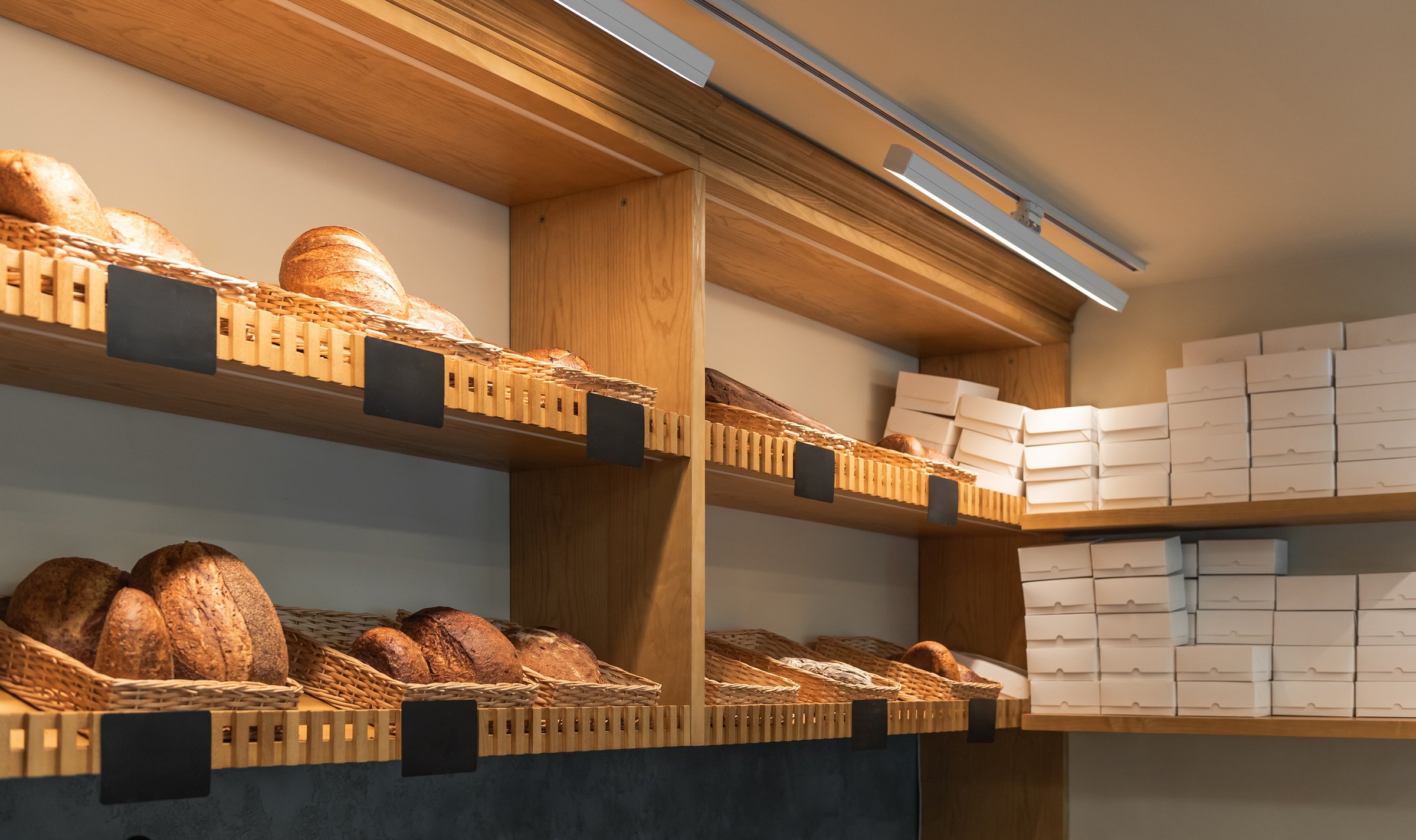 Artisanal bread loaves sit in wicker baskets on wooden shelves to the left. To the right, tall stacks of plain white takeout boxes are stored on shelves.