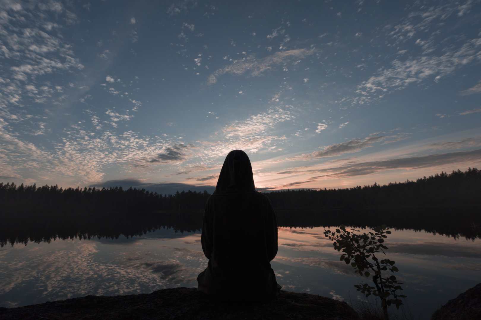 Silhouette of a hooded woman on the shore of the lake. The sunset sky is reflected in the mirrored surface of the water