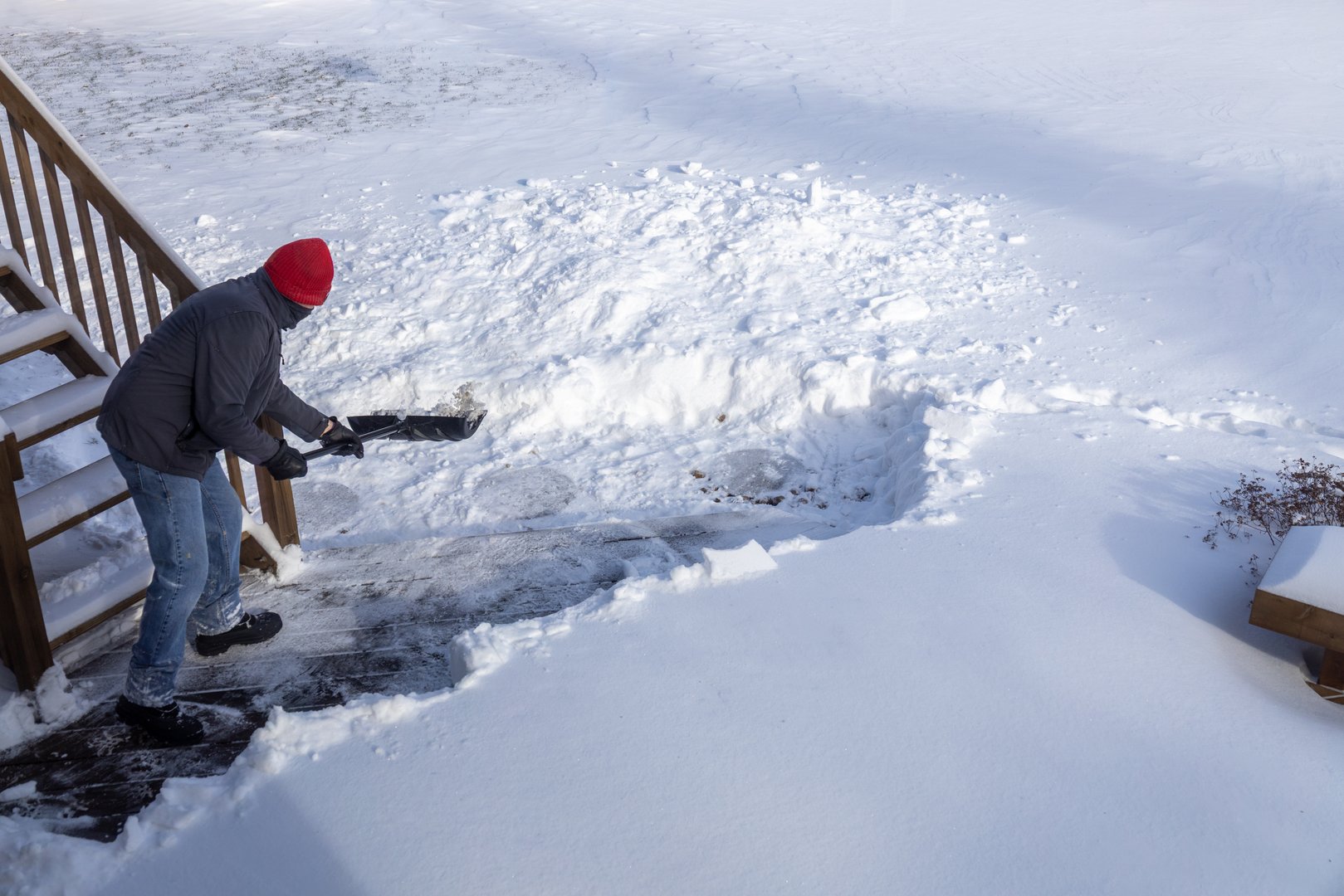 This image shows an unrecognizable adult man shoveling deep snow off of a wooden deck, following a blizzard.
