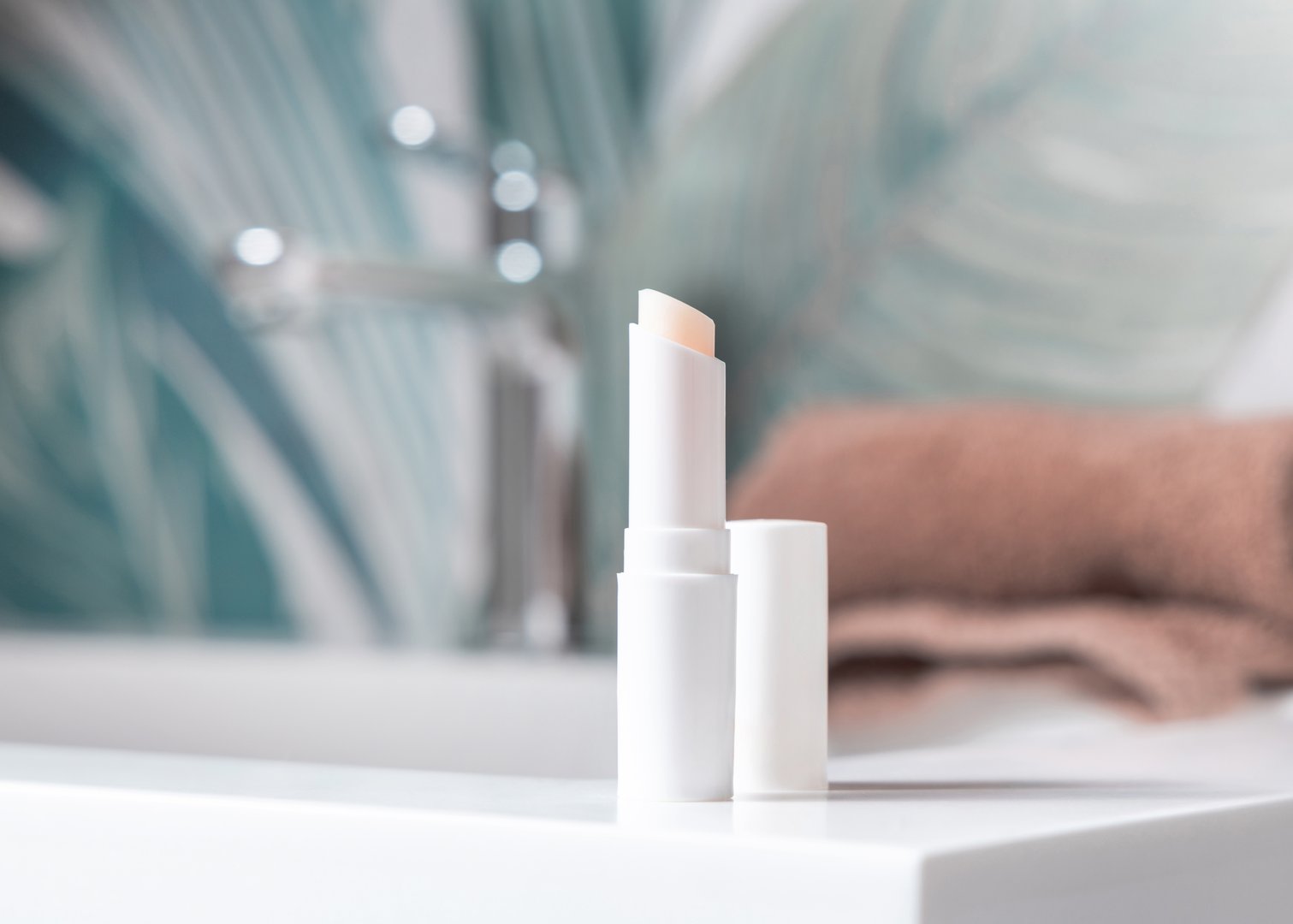 Opened white blank lip balm stick with a lid near brown towel on basin in green tropical bathroom close up, packaging mockup with copy space. Lifestile scene with ubranded natural lipcare product
