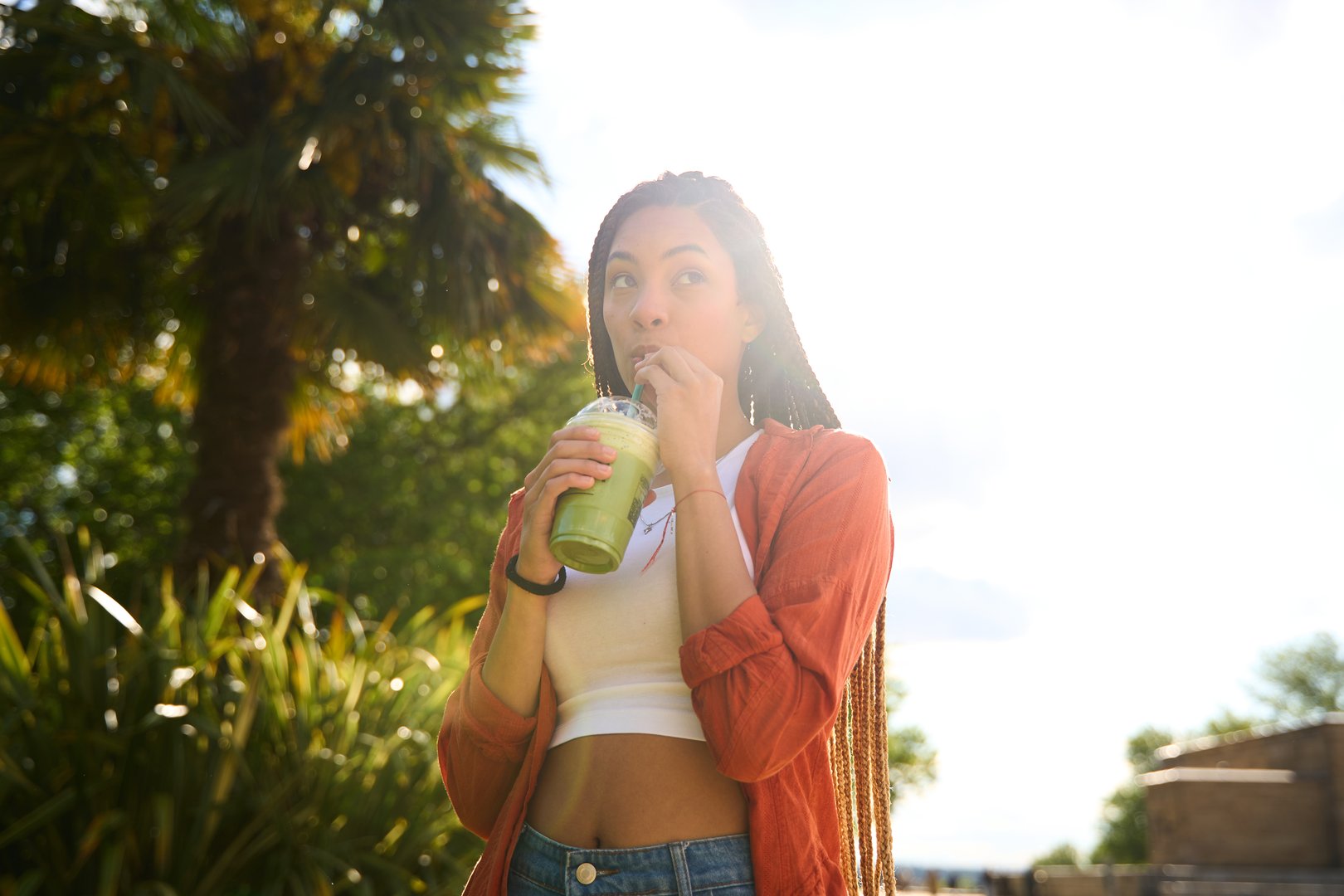 Young woman with long braids drinking a green smoothie outdoors in a park during a sunny summer day