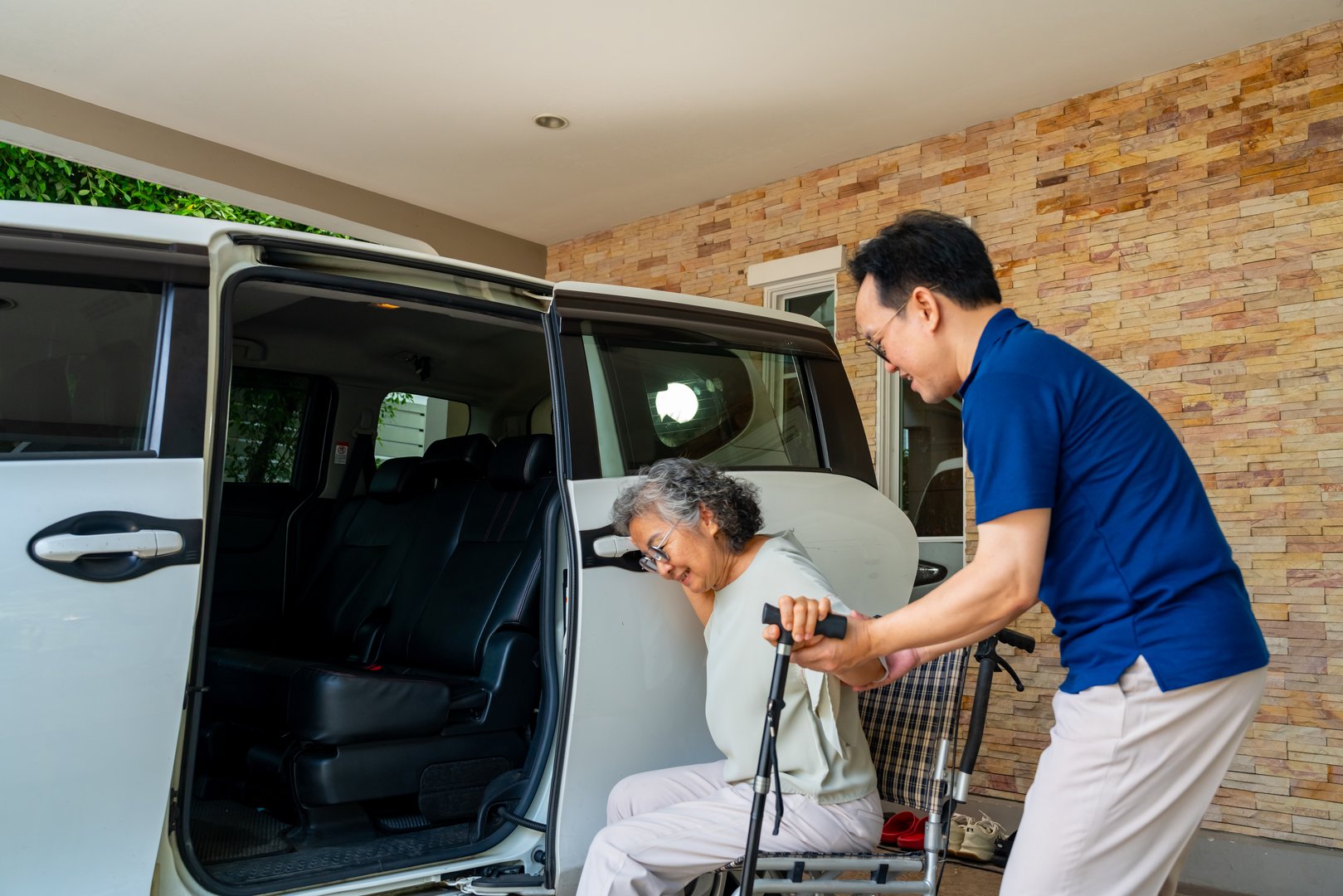 Asian man caregiver or taxi driver helping senior woman patient get in the car to get to medical appointments or healthcare therapy sessions at hospital. Assisted elderly patient transportation services