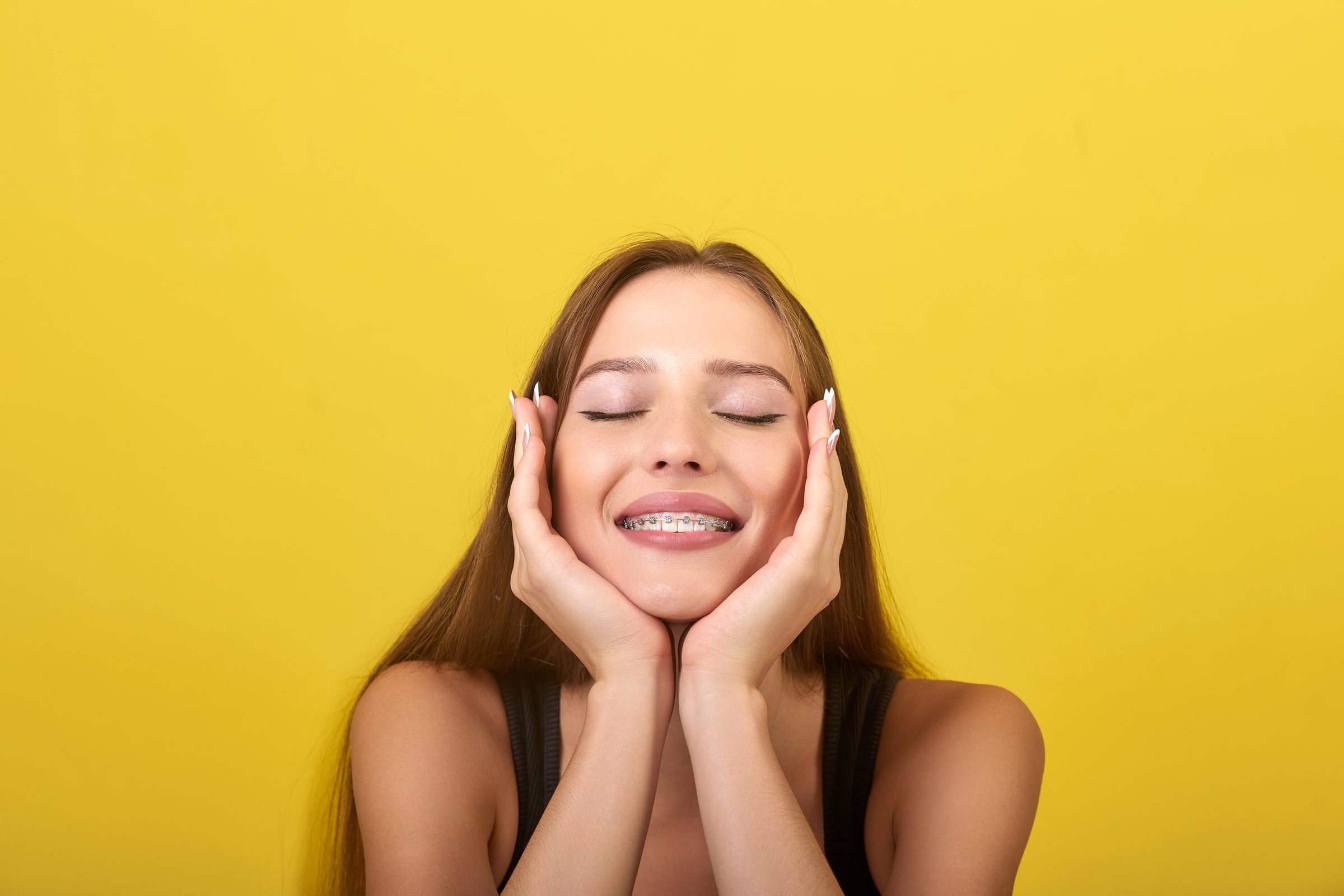 The image shows a young woman with long brown hair and braces on her teeth. She is wearing a black tank top and has her hands on her face. The background is a bright yellow color.