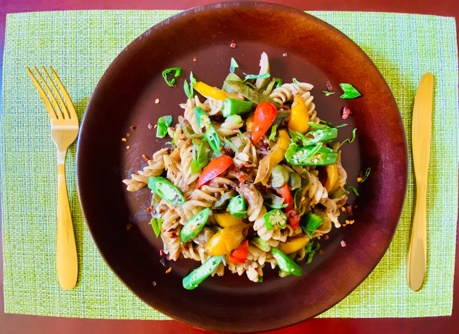 Pasta salad with colorful vegetables on a brown plate, set on a green placemat with gold cutlery.