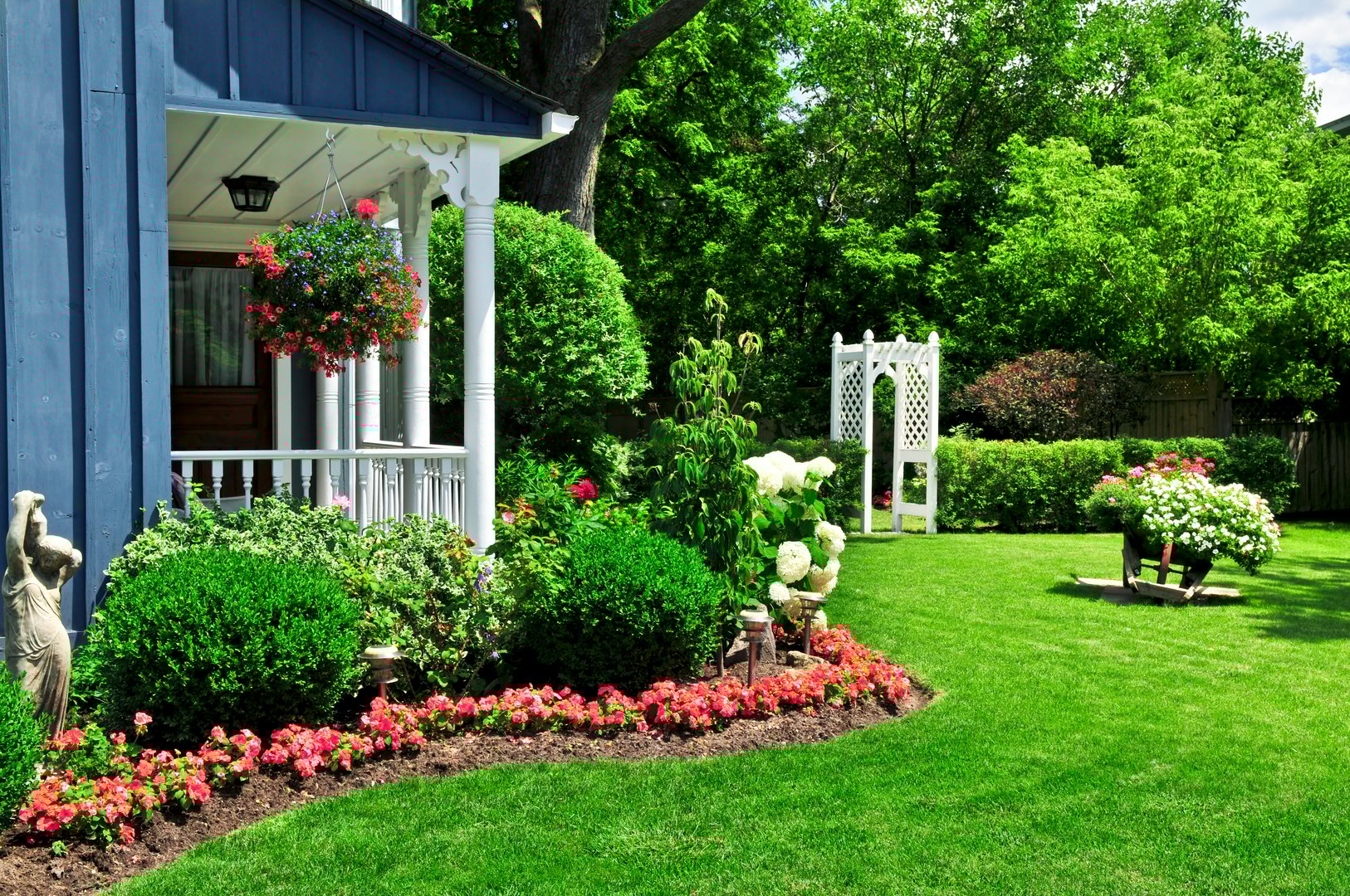 House with a porch, garden with pink flowers and bushes, white trellis, and statues in a well-manicured lawn.
