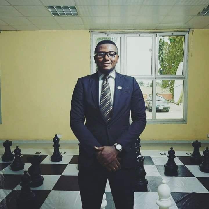 Man in suit stands on large chessboard in a room with yellow walls and a window, surrounded by big black chess pieces.