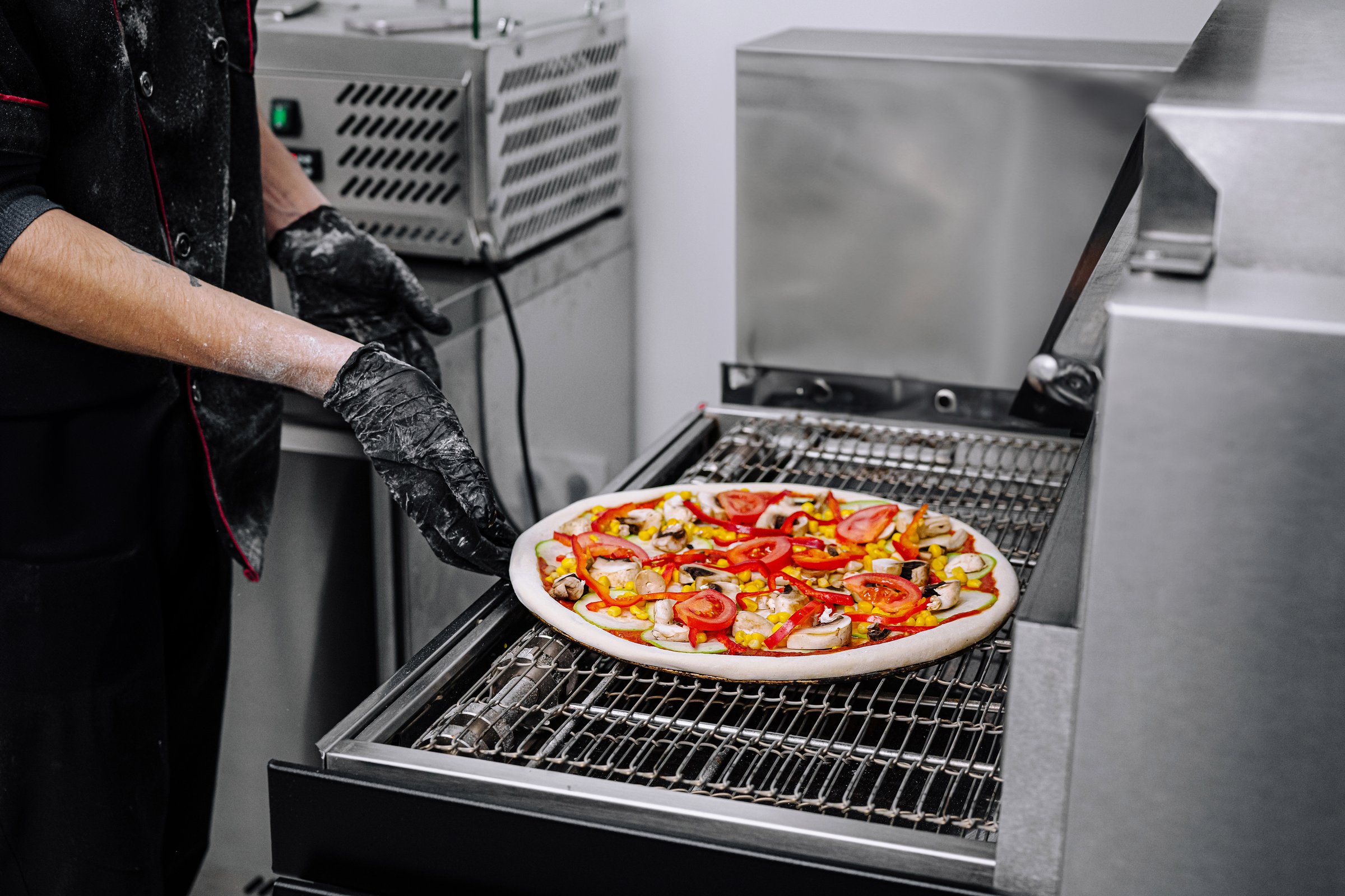 A chef wearing gloves is placing a pizza topped with fresh vegetables onto a grill in a contemporary kitchen. The vibrant colors of the ingredients stand out.