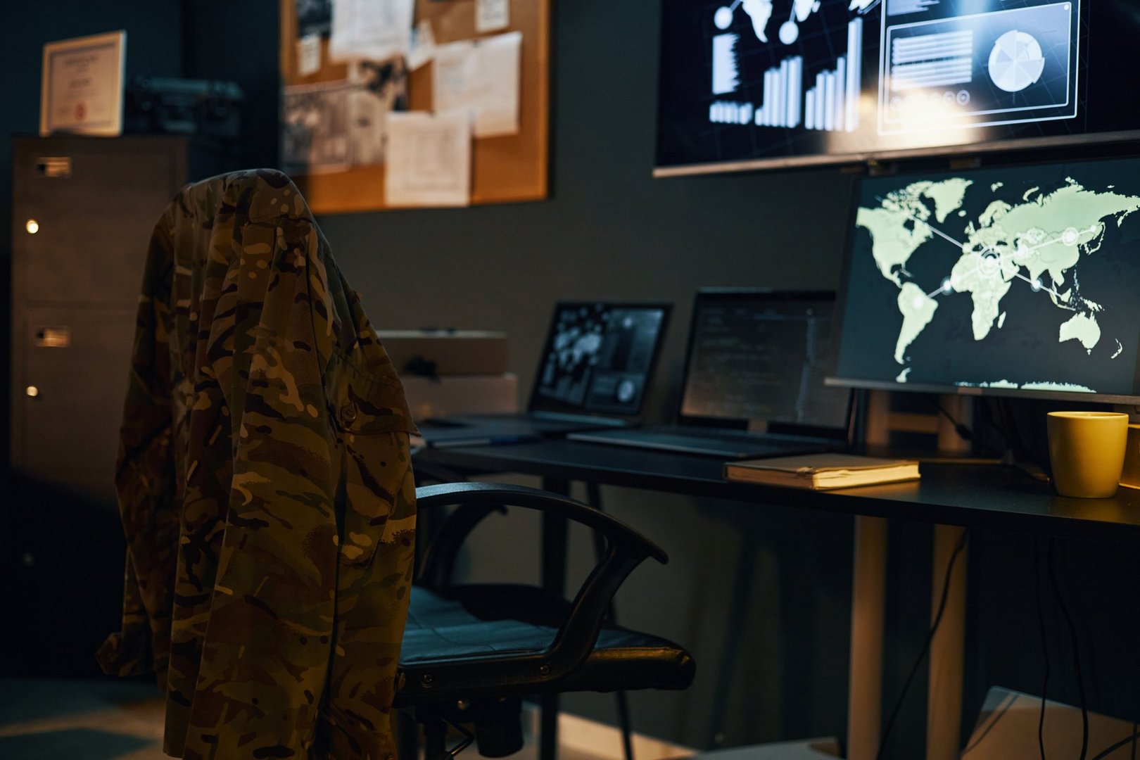 Command center featuring multiple computer screens with real-time data and maps, camouflage jacket on chair, creating focus on crucial operations and communications