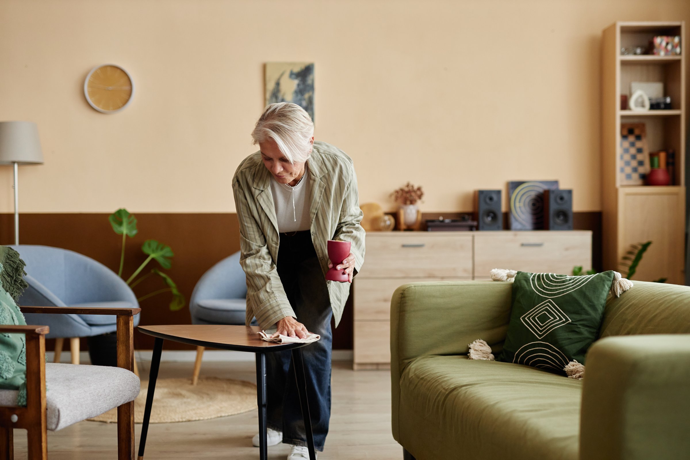 Full length portrait of elegant mature woman cleaning home and dusting coffee table in cozy living room copy space