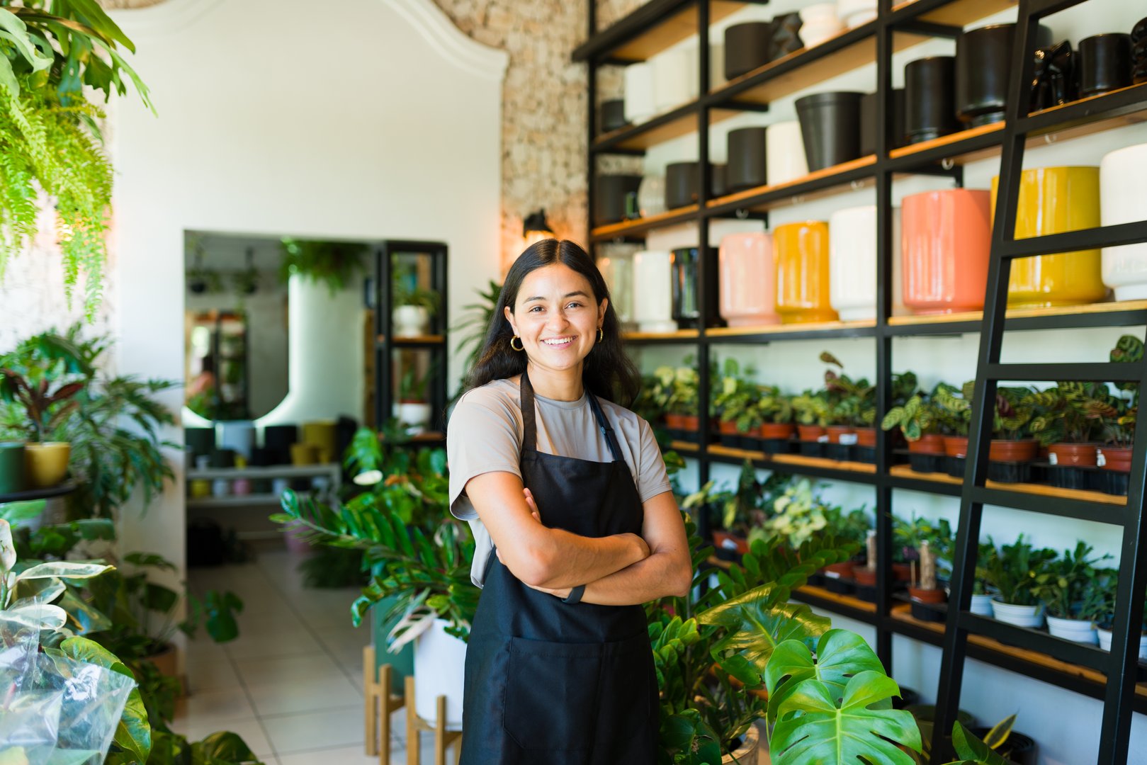 Happy young hispanic woman entrepreneur standing with arms crossed and smiling in her plant shop