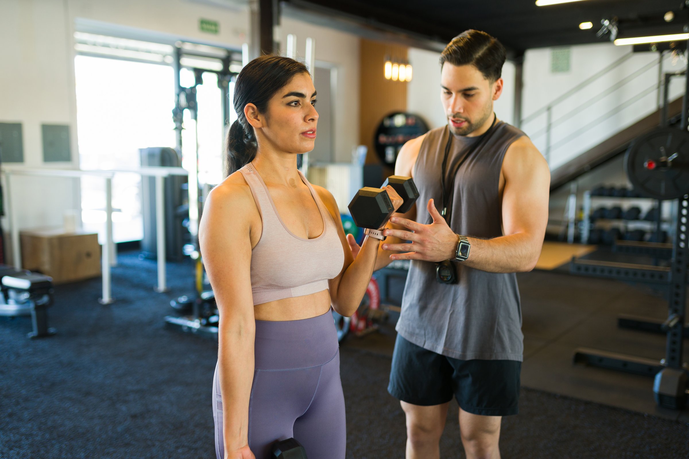 Female client performing bicep curls with trainer assistance, targeting arm muscles during strength training session in contemporary fitness center