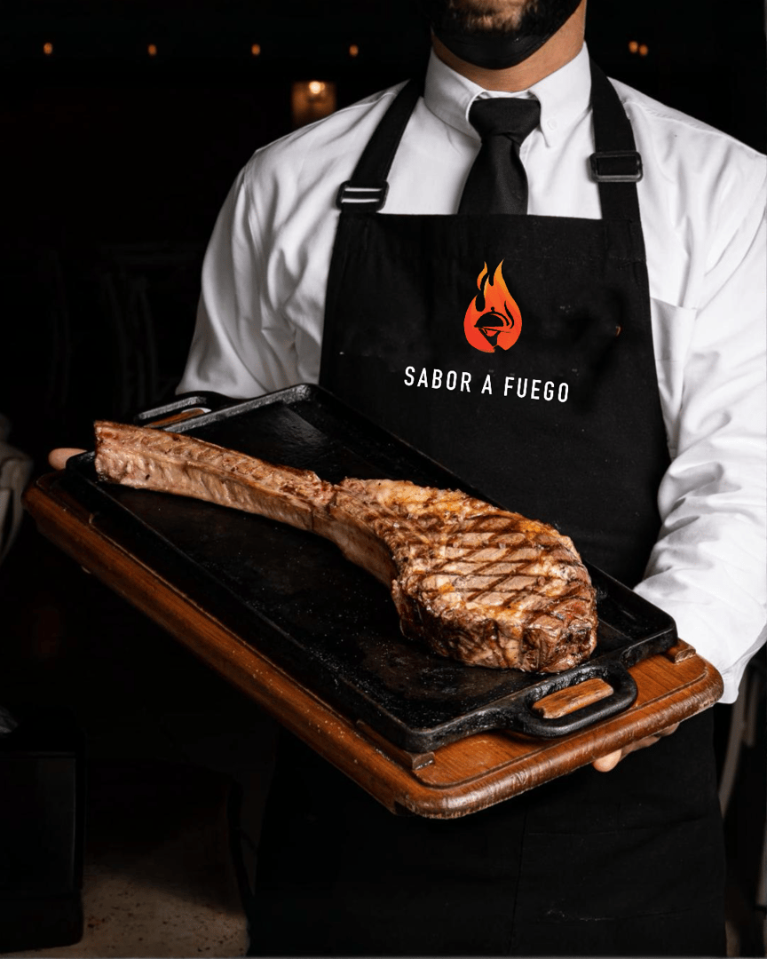 A waiter in a black apron holds a tray with a grilled tomahawk steak on a wooden board, branded with "Sabor A Fuego."