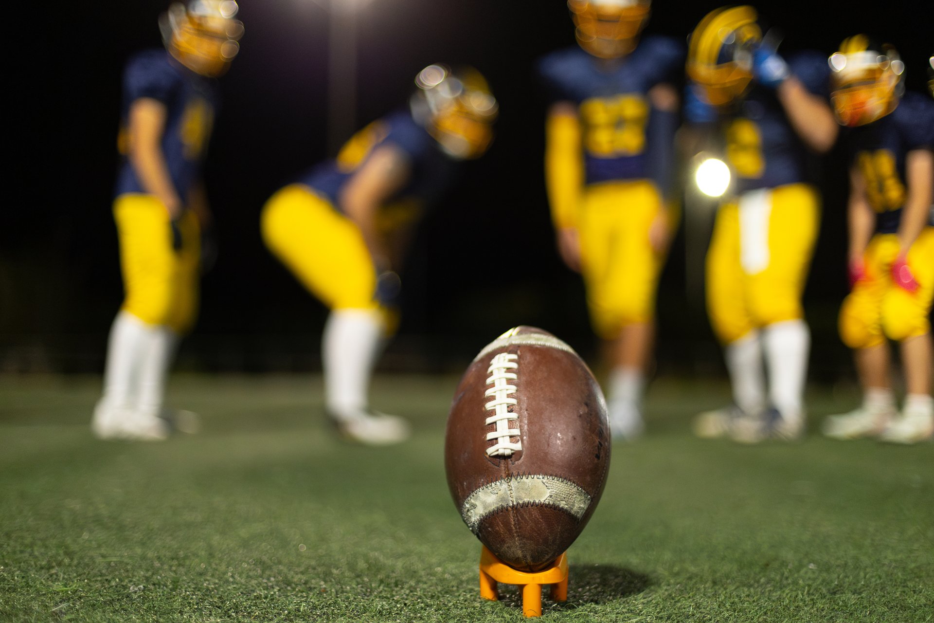American football and kicking tee on artificial turf field with blurred players in uniforms standing in background at night