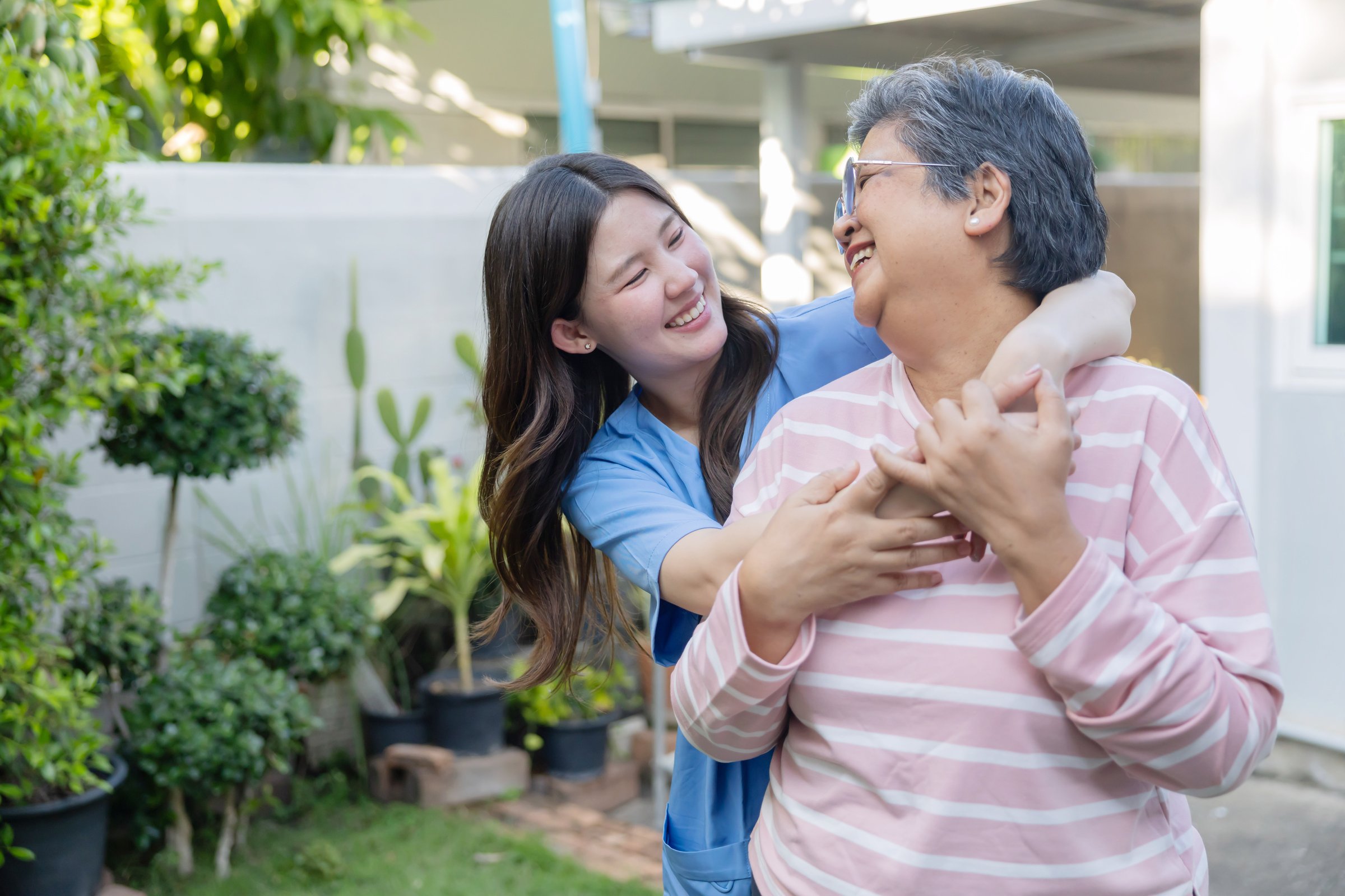 Two women share a joyful moment in a garden. One woman hugs the other from behind