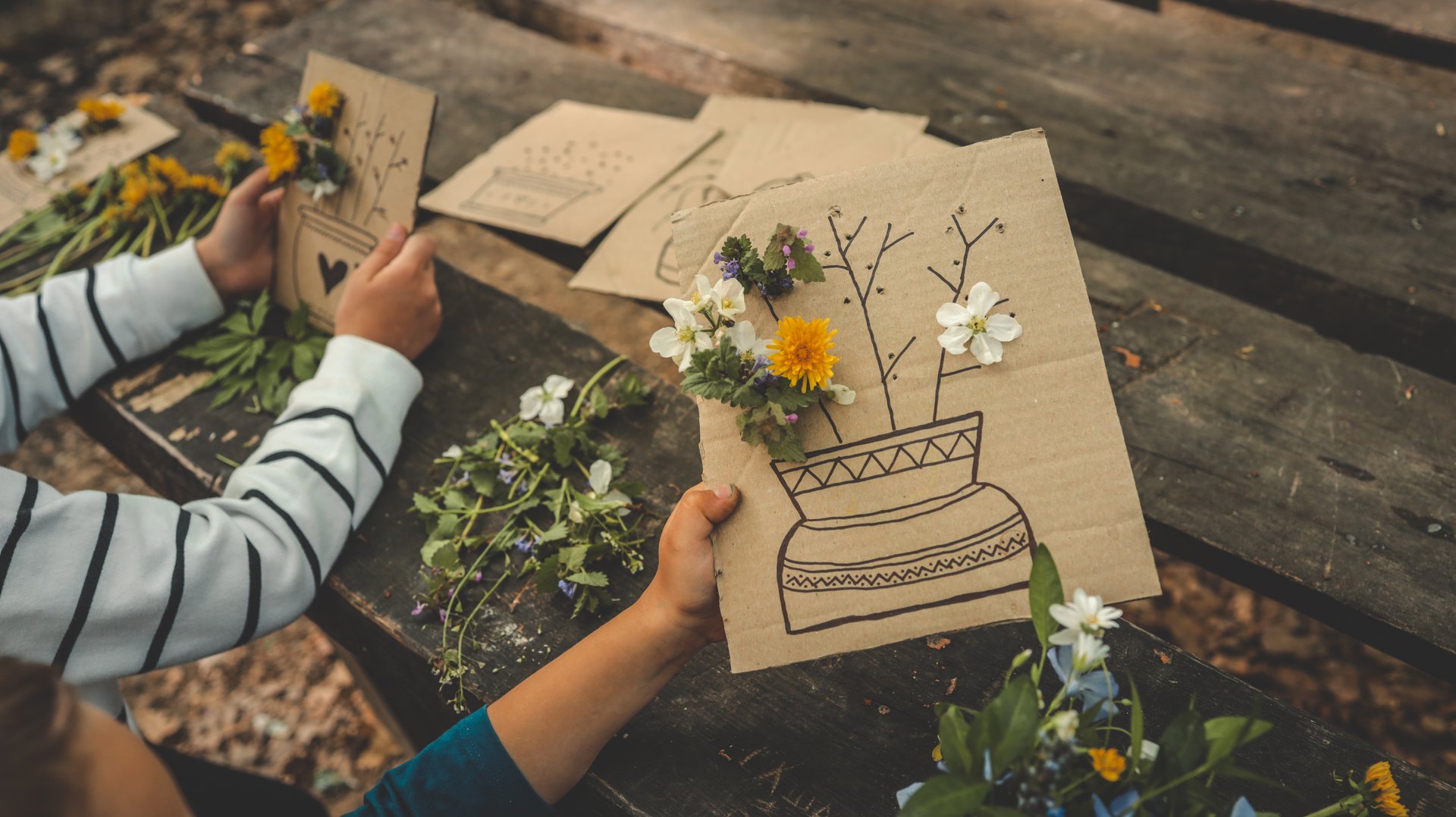 Children creating handmade flower crafts using cardboard, drawing vases and attaching real spring blossoms. Creative outdoor activity on a rustic wooden table in the forest.