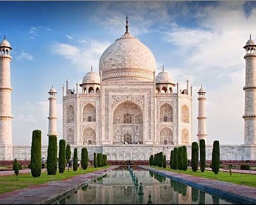 The Taj Mahal with its white marble dome and minarets, reflected in a long reflecting pool, surrounded by manicured gardens.