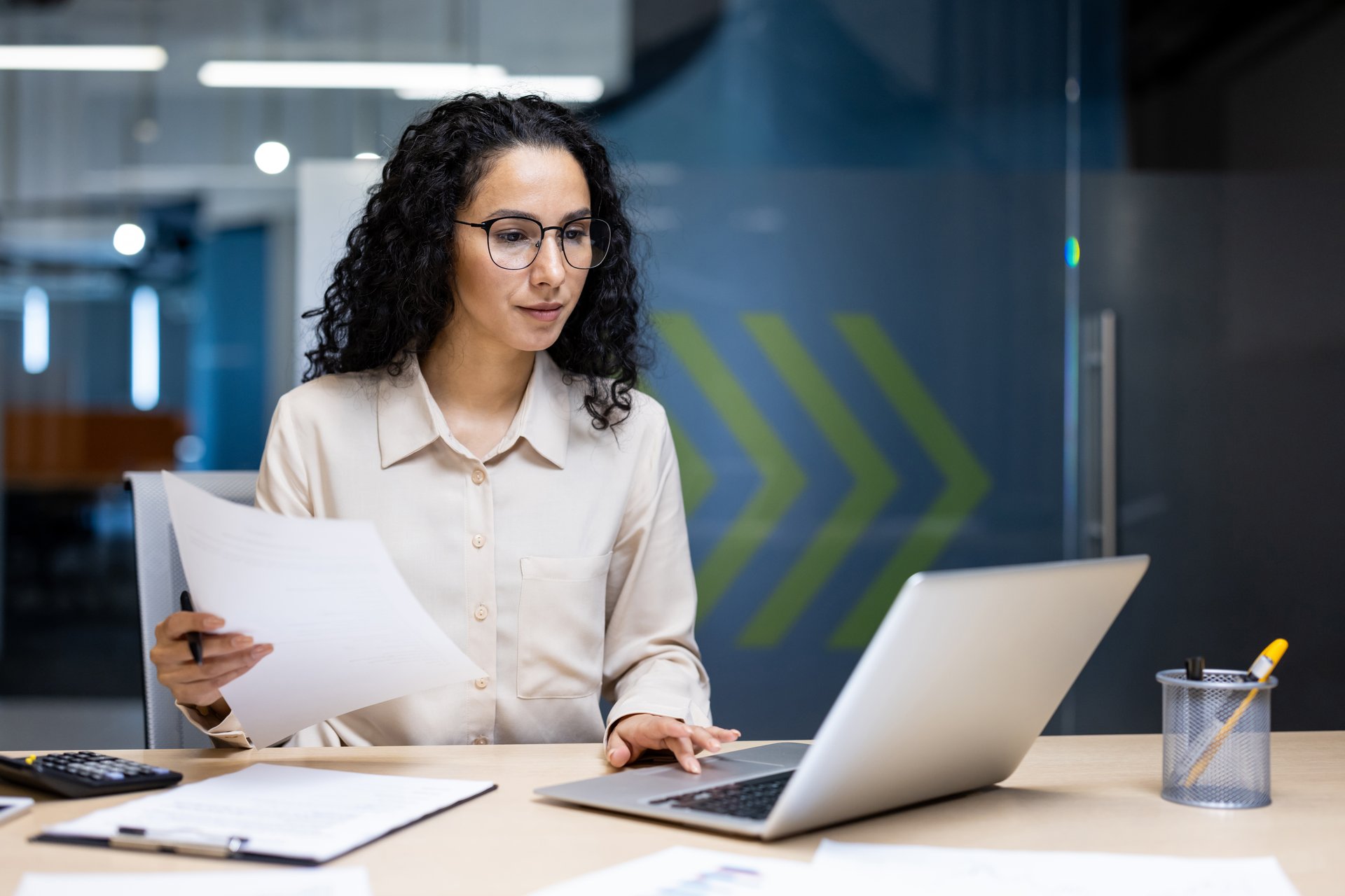 Serious and pensive business woman behind paper work inside office, female financier worker thinks about contracts and reports with charts and graphs, hispanic successful woman uses laptop at work