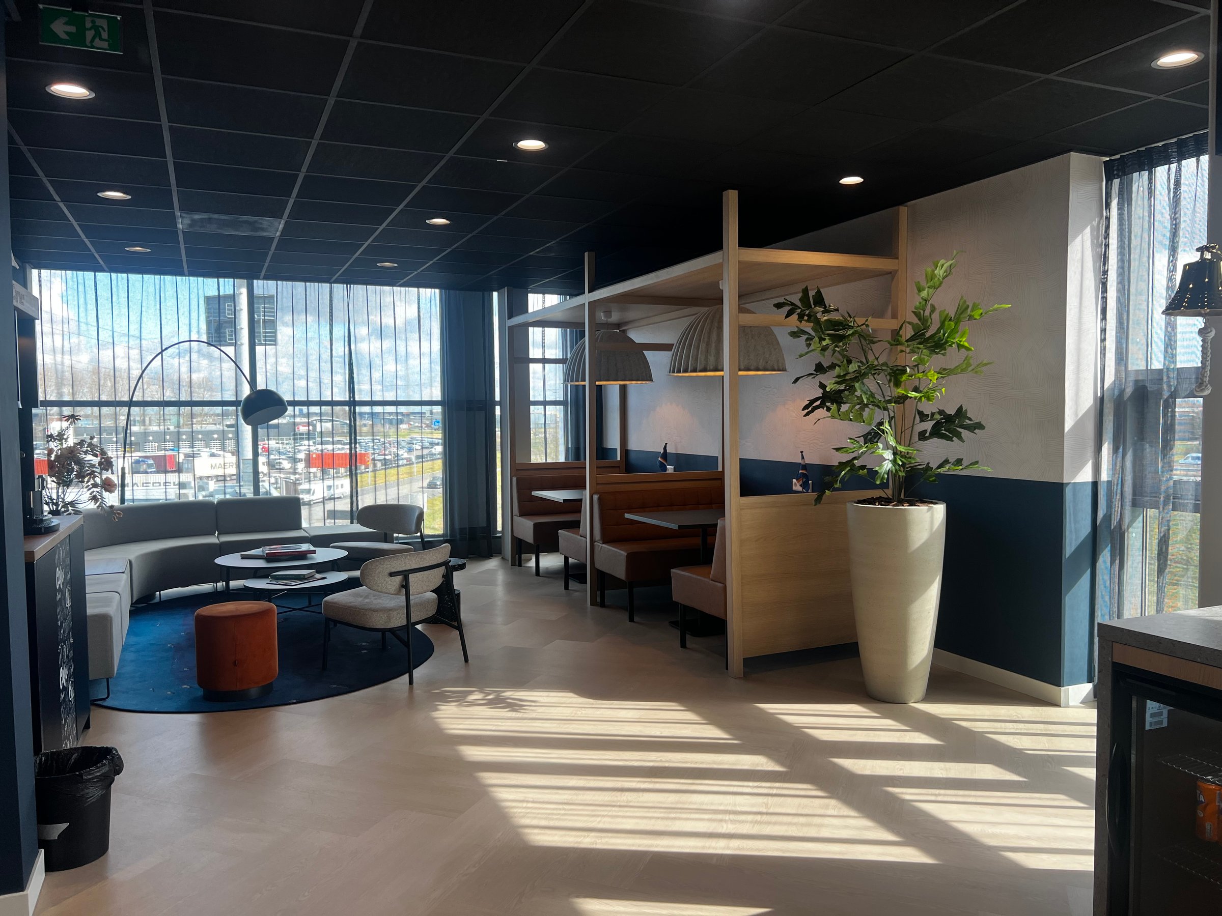 Modern office lounge with seating area, tables, plants, and large windows letting in natural light.