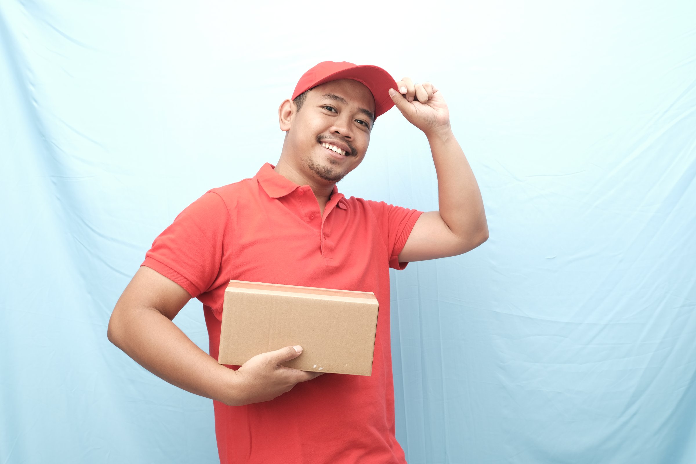 Friendly delivery worker in a red uniform, holding a parcel and tipping his cap with a smile against a light blue background.