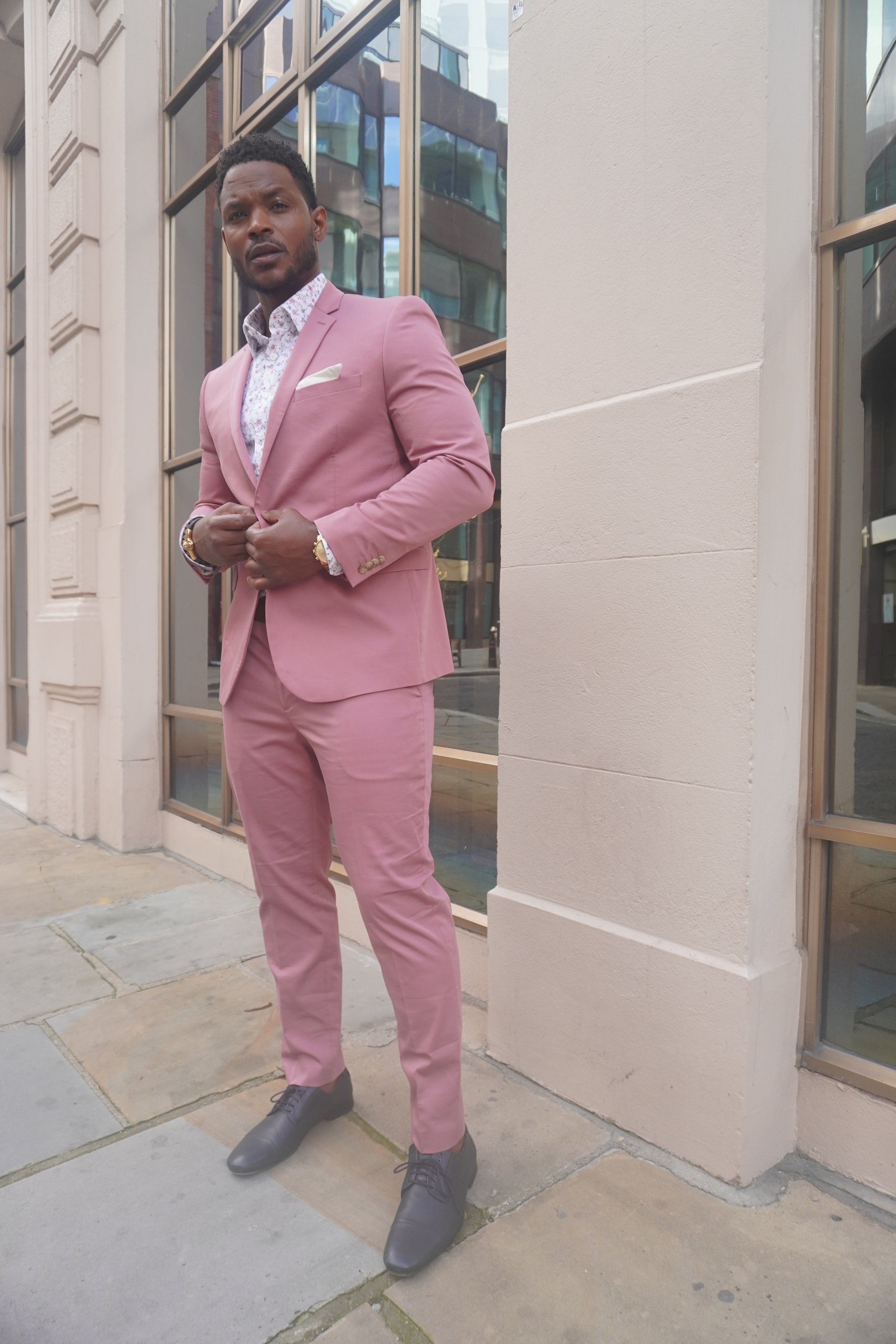 Man in a pink suit and patterned shirt stands confidently on a city street, near a building with large windows.