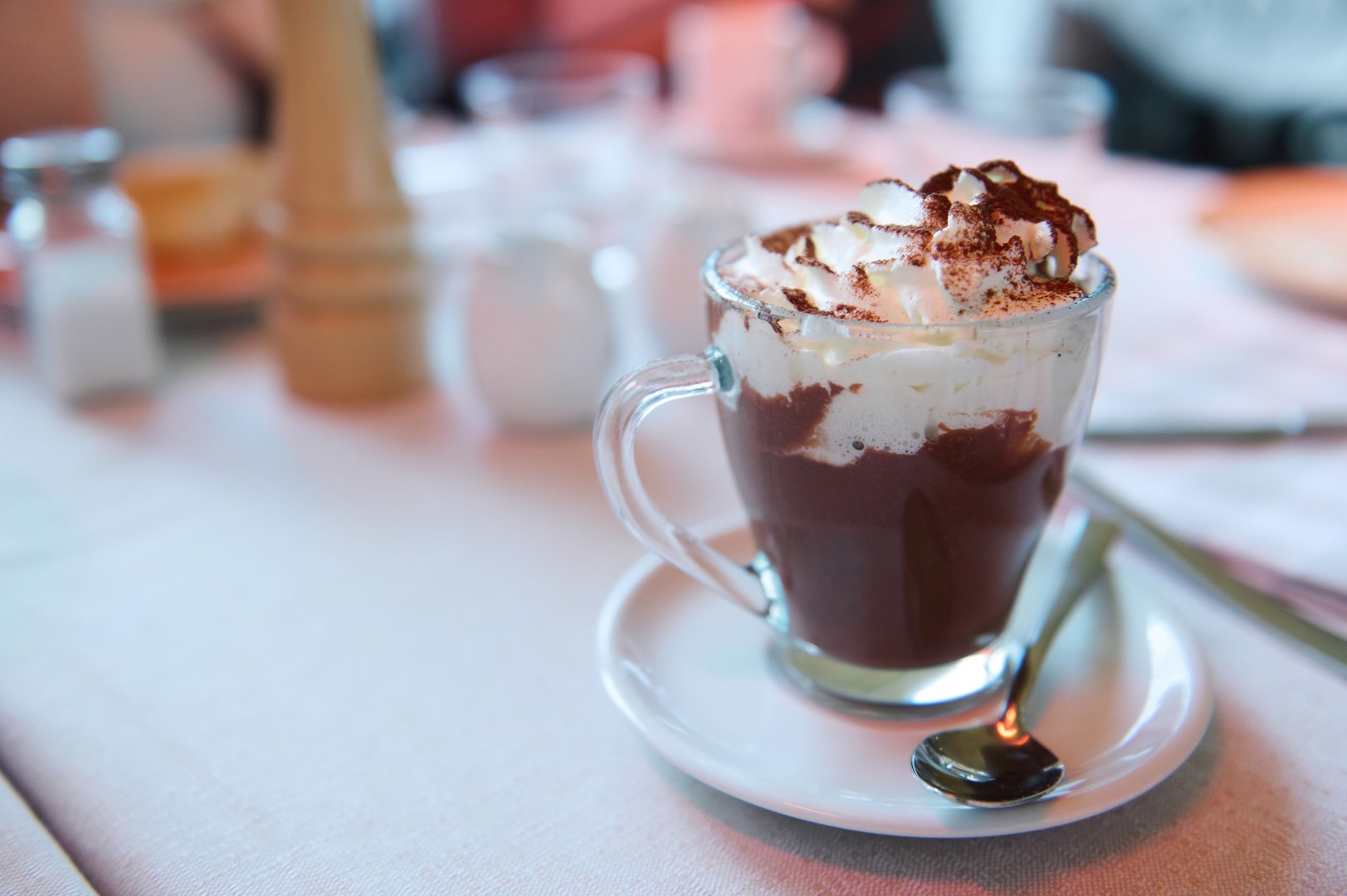 Close-up of a glass mug filled with hot chocolate, topped with whipped cream and cocoa powder on a table setting in a cozy cafe.