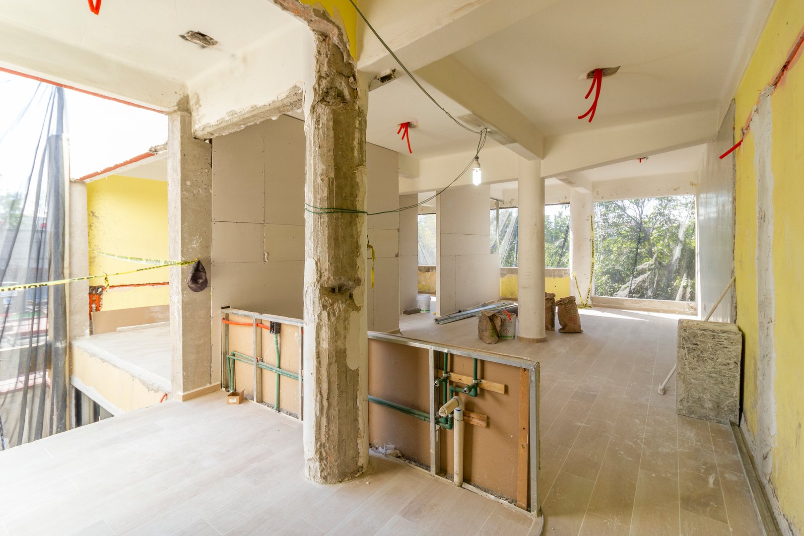 Room under construction with exposed walls, ceiling, and wiring. Sunshine filters through large windows, illuminating the space.